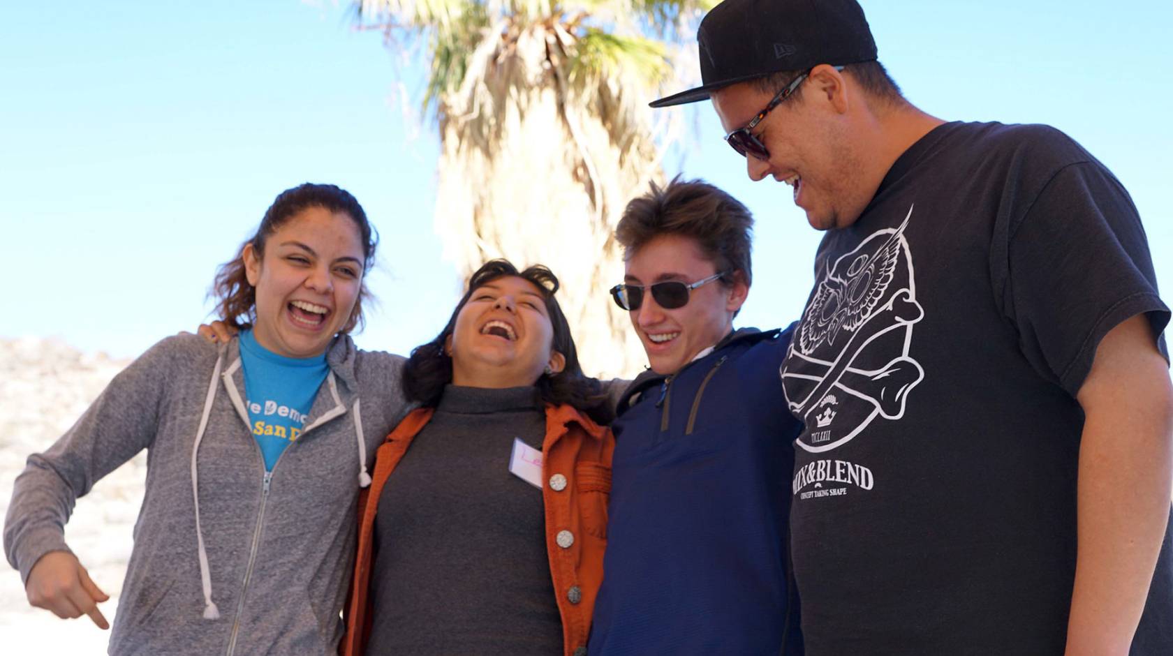 Four students with arms around each other laugh and smile, with a palm tree in the background