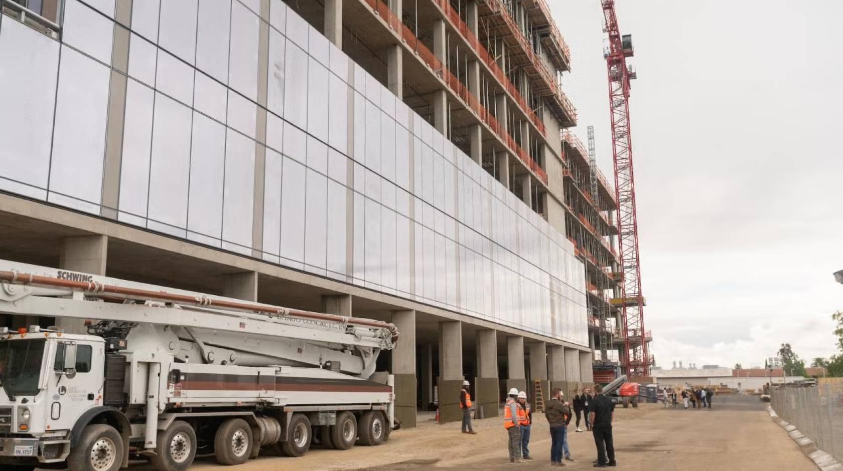 A building  with numerous glass panels under construction on UC Davis' Aggie Square with a construction crew talking below it next to a truck with a crane