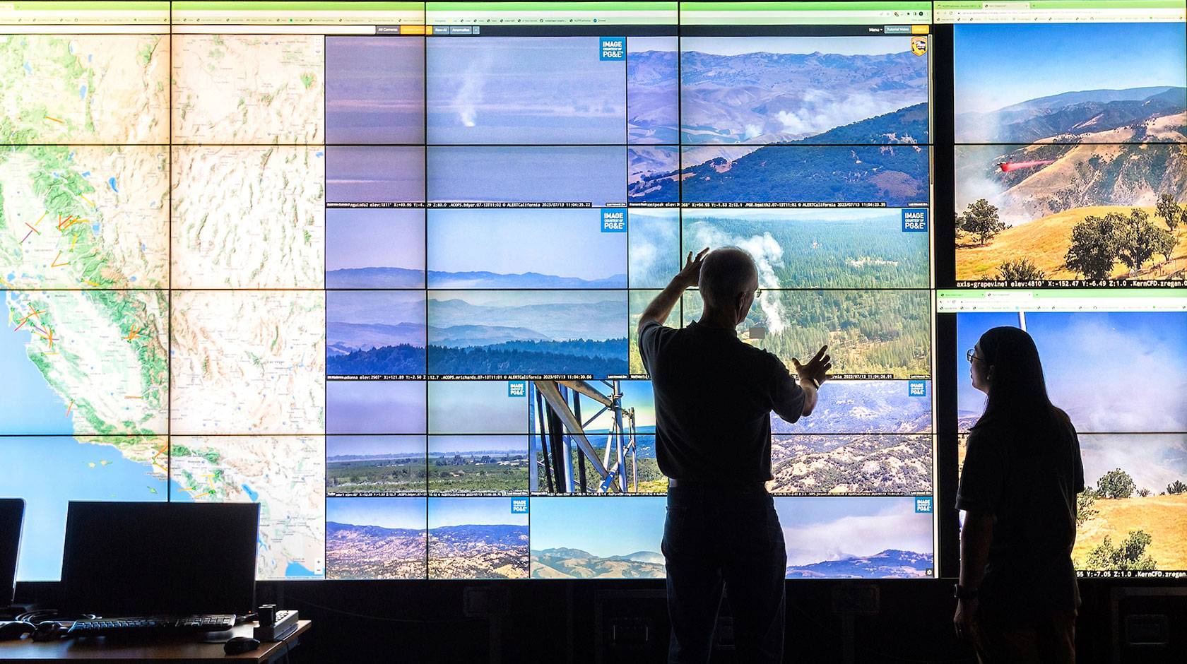 Two people stand in front of a wall of screens showing a grid of footage of natural landscapes, a map of California and some smoke from a wildfire 