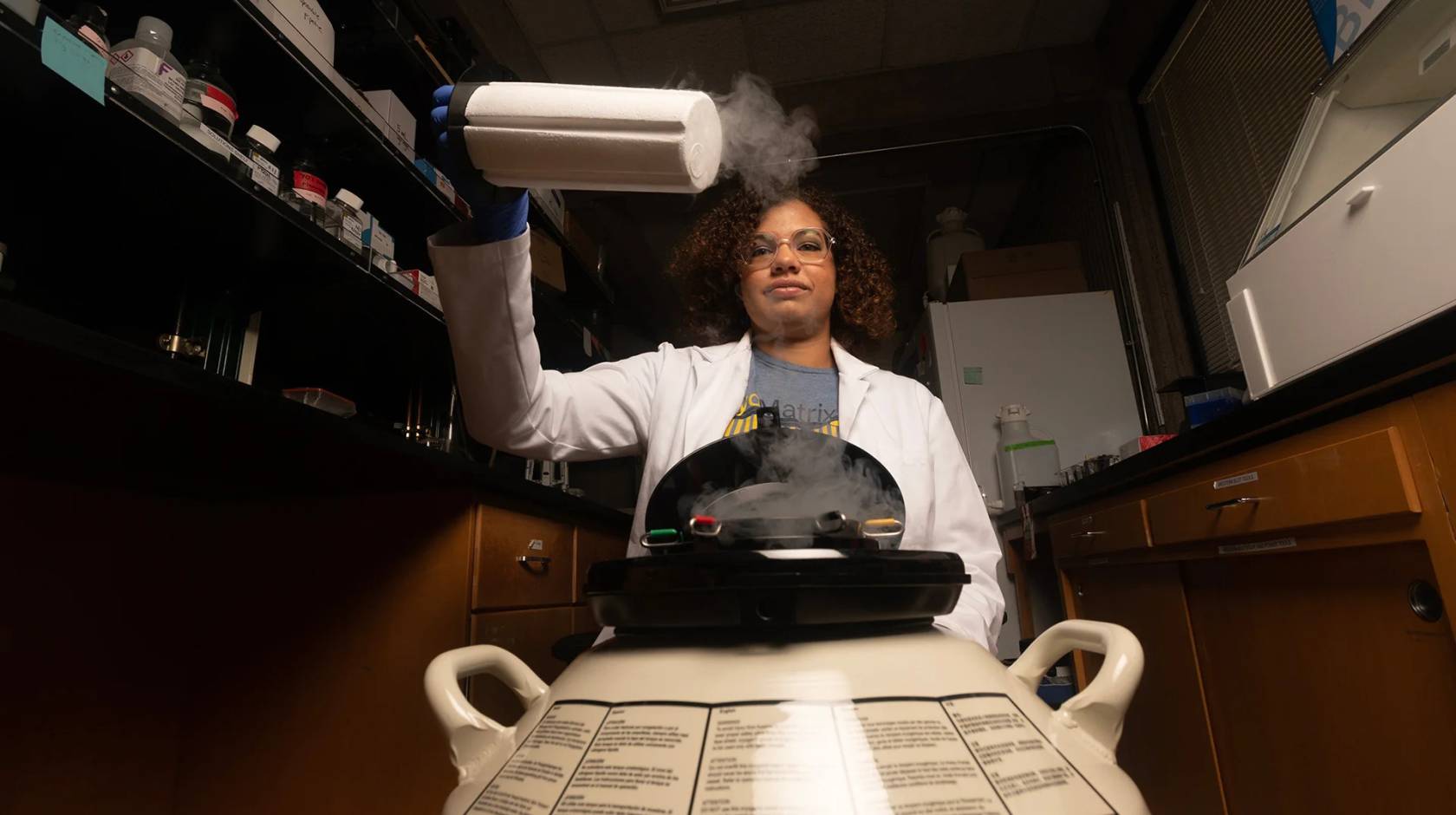 A young woman graduate student with curly hair pulls out a nitrogen storage cylinder and holds it over a piece of equipment