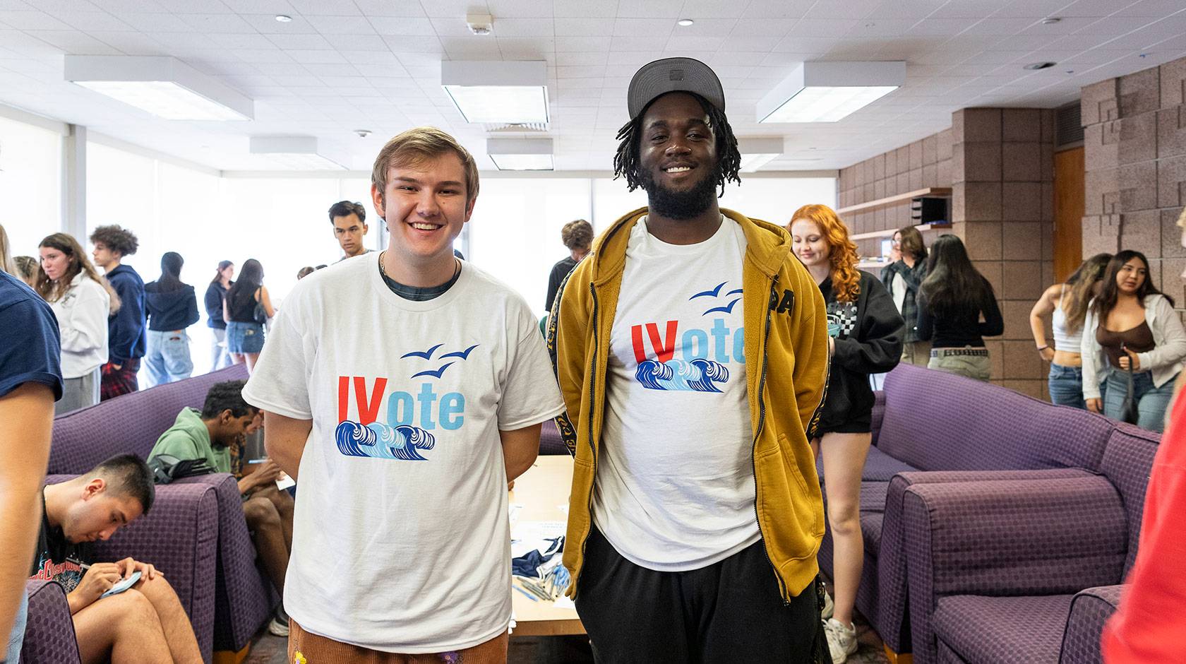 Two UC Santa Barbara students face the camera and smile, wearing t-shirts reading "IV Votes", standing in a busy student lounge.