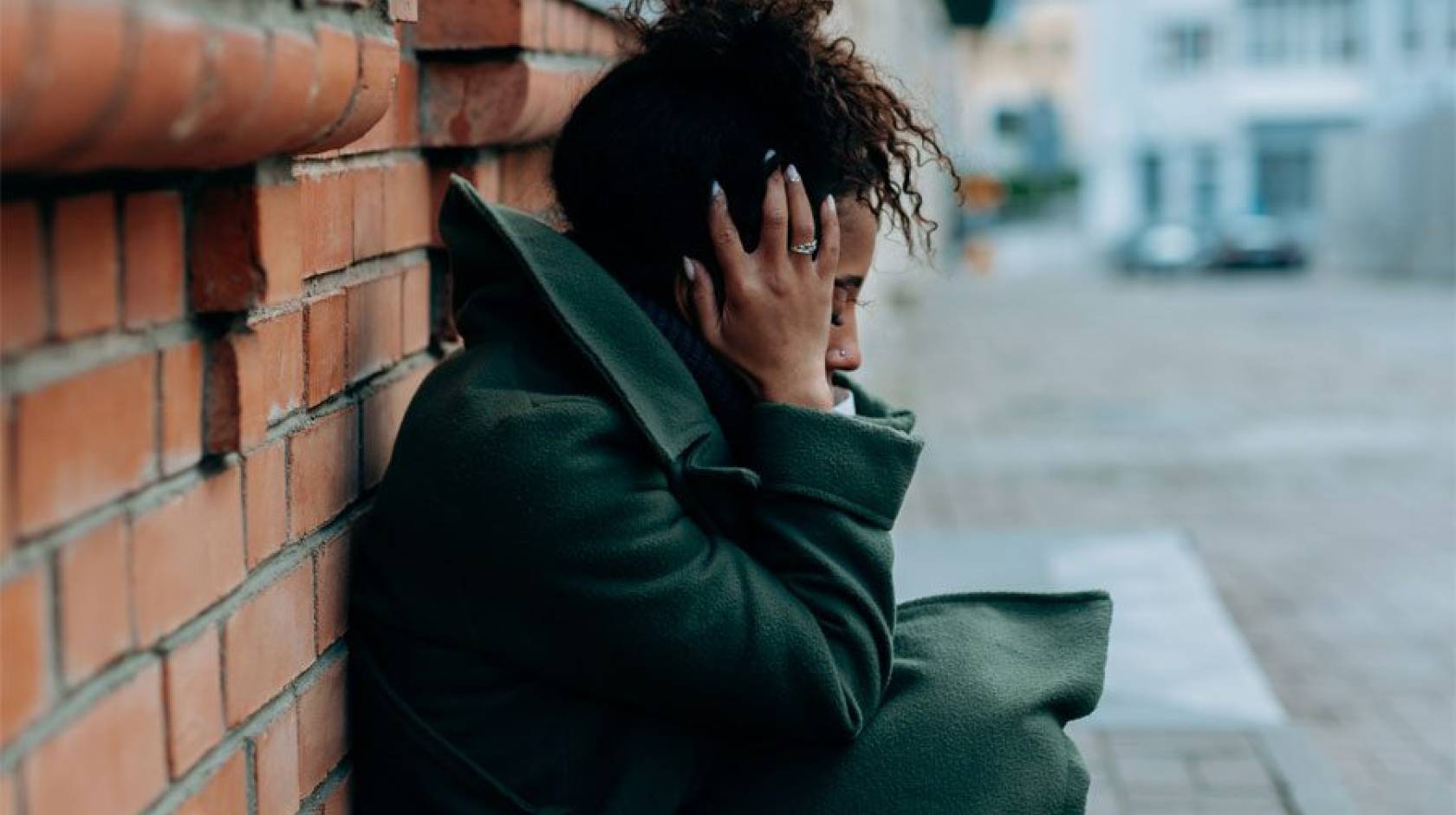 A young Black woman crouches against a brick wall holding her head