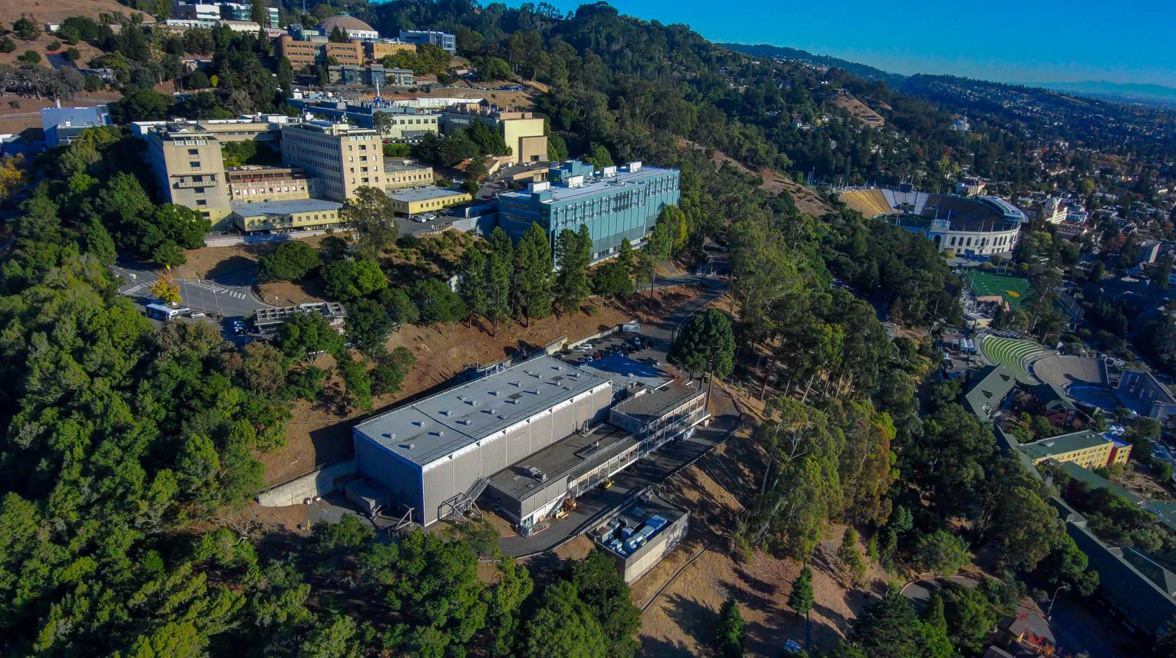 A number of buildings on a tree-covered hillside, including Berkeley Lab, seen from an aerial view on a clear day, Bay in the background