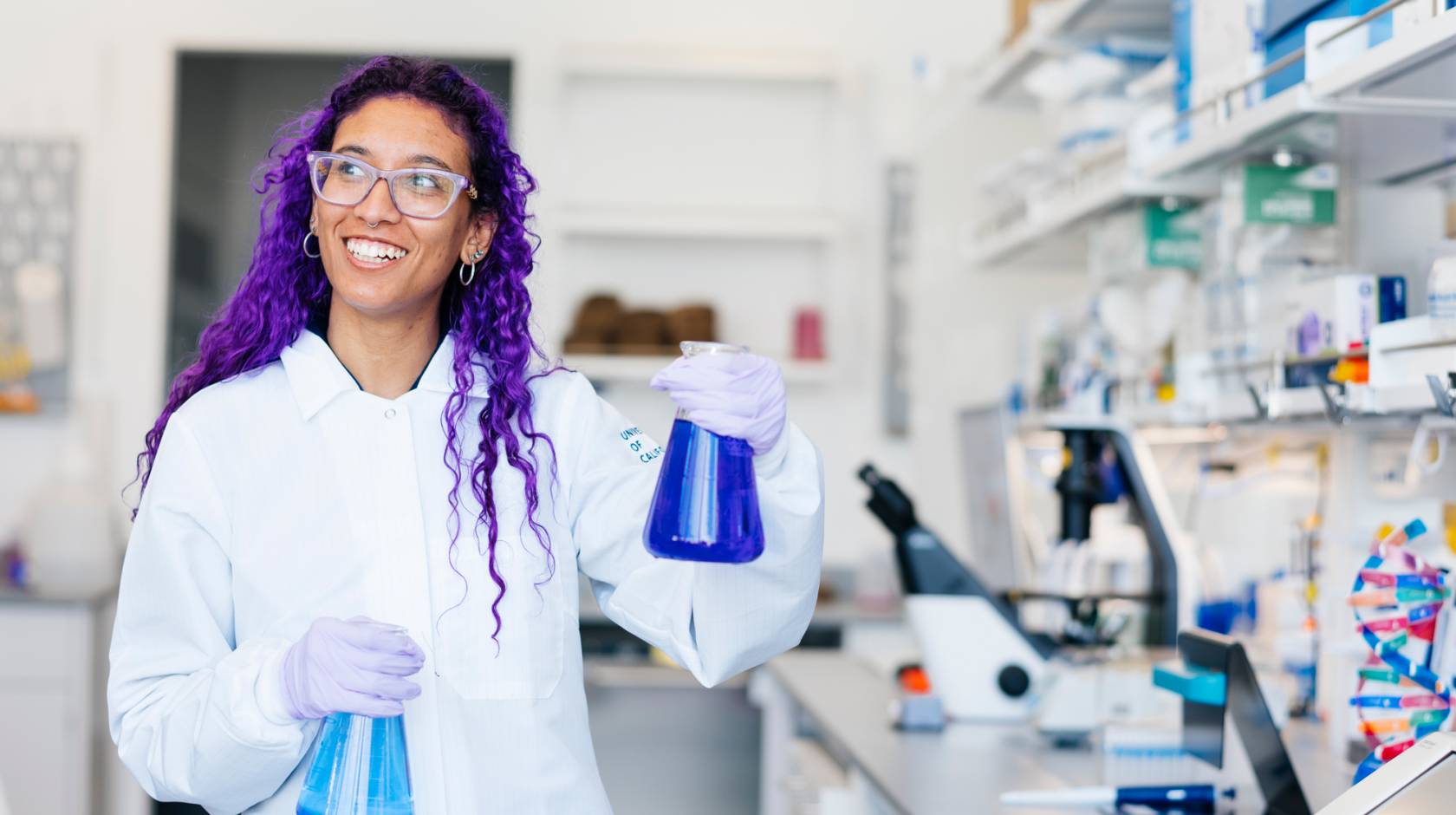 A woman with two flasks of blue chemicals and long purple hair in PPE and a lab coat smiles at a lab table