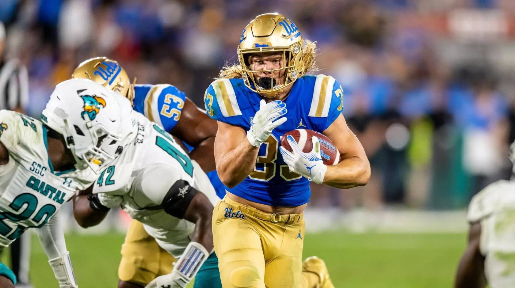 A player in UCLA blue and gold carries the ball and runs by players in white uniforms