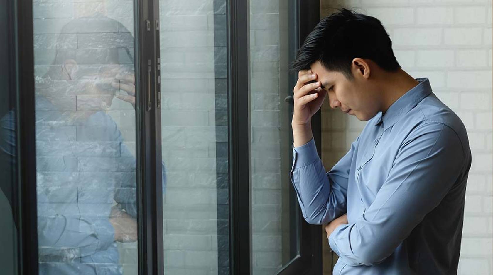 Young Asian man holds head in hand in front of his reflection at work