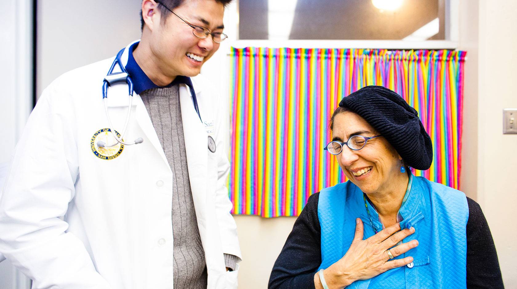 A doctor in a white coat laughs with a patient, who is smiling and holding her hand over her heart.