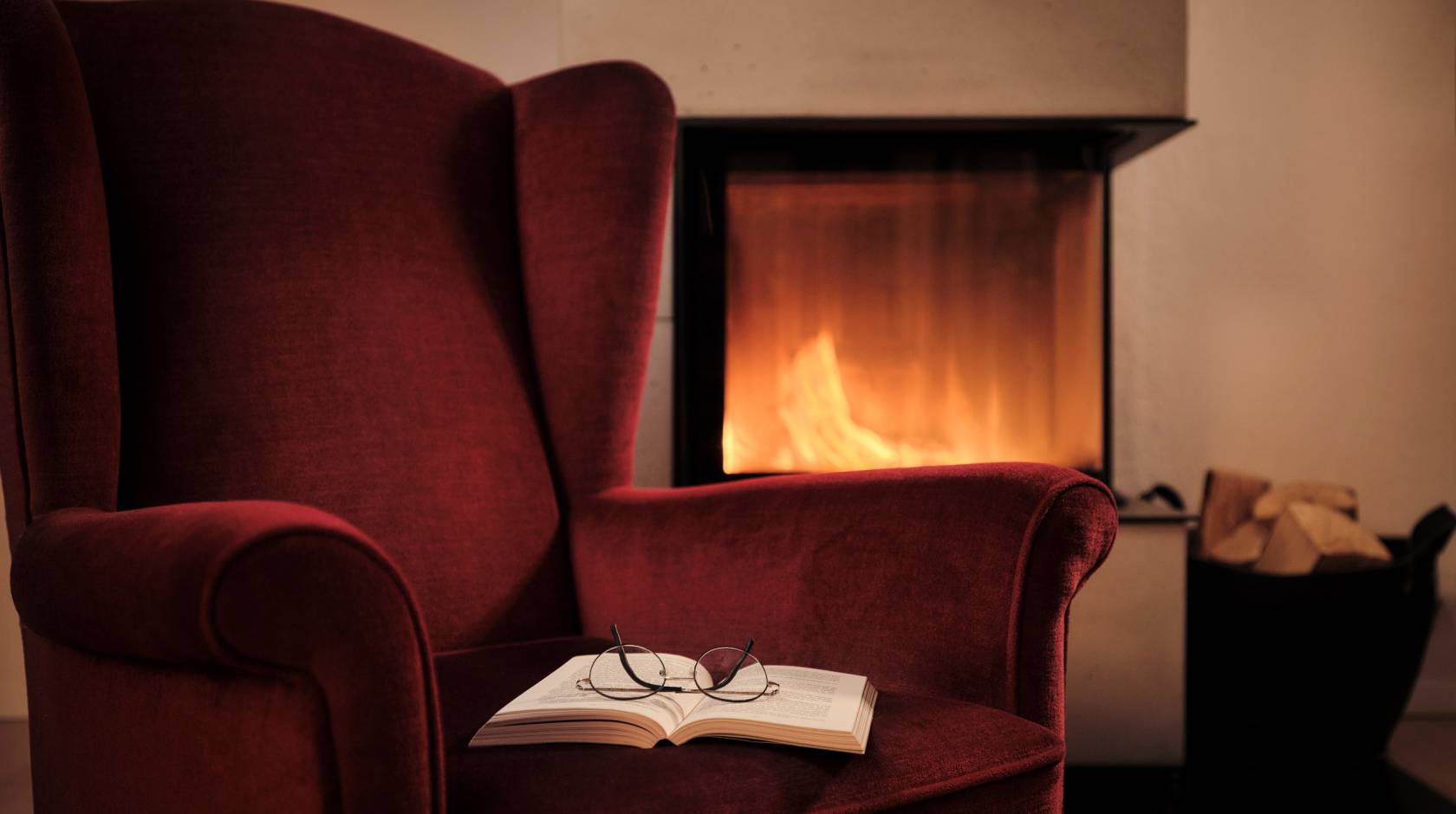 A deep red arm chair with an open book with glasses on top of it next to a fireplace