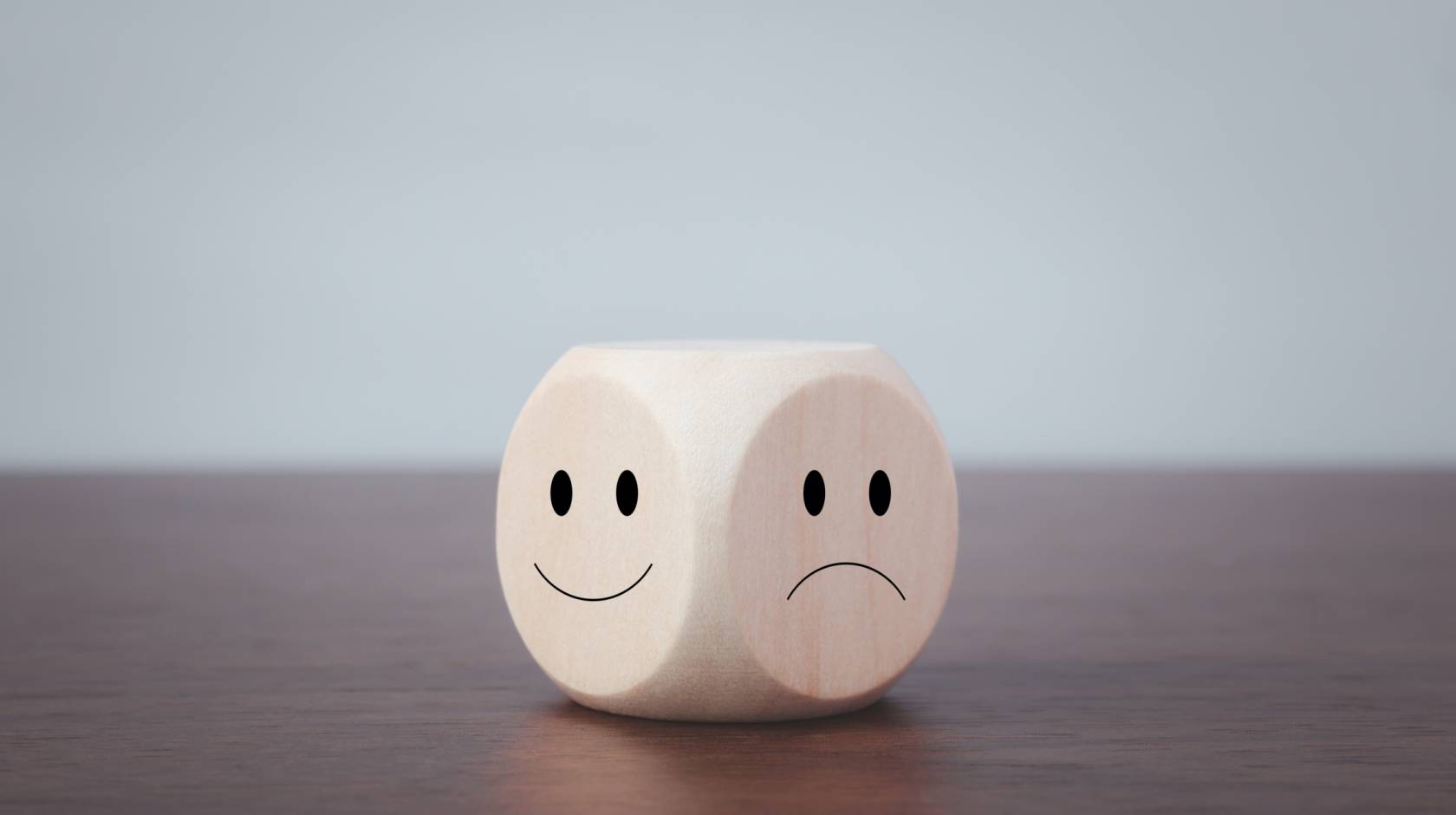 A 6-sided die on a wooden table top. Of the 2 faces visible, one has a happy face and the other has a sad face drawn on it