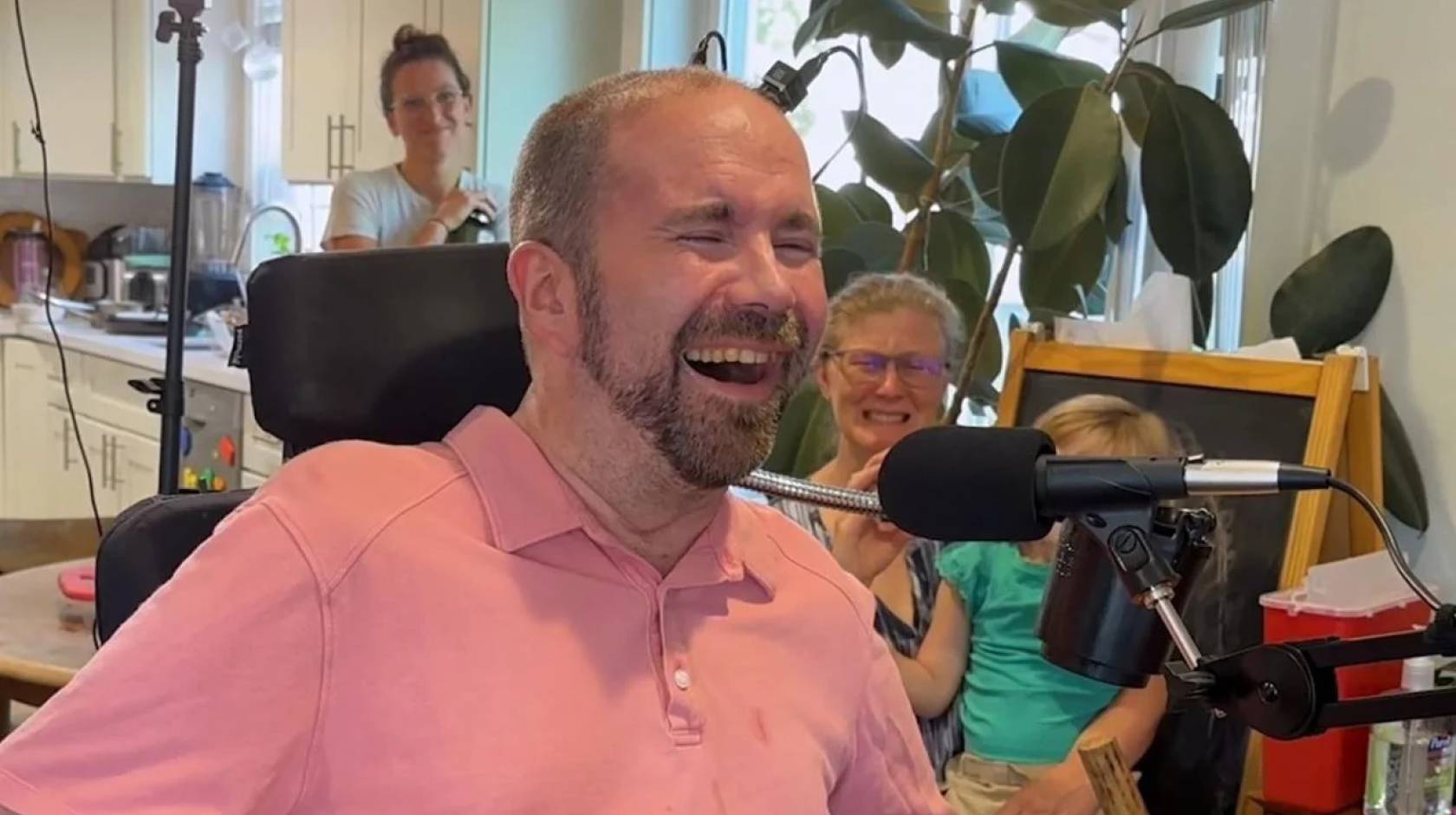 A man with a beard and a pink shirt in his living room grinning, overjoyed in front of a microphone, while his family watches