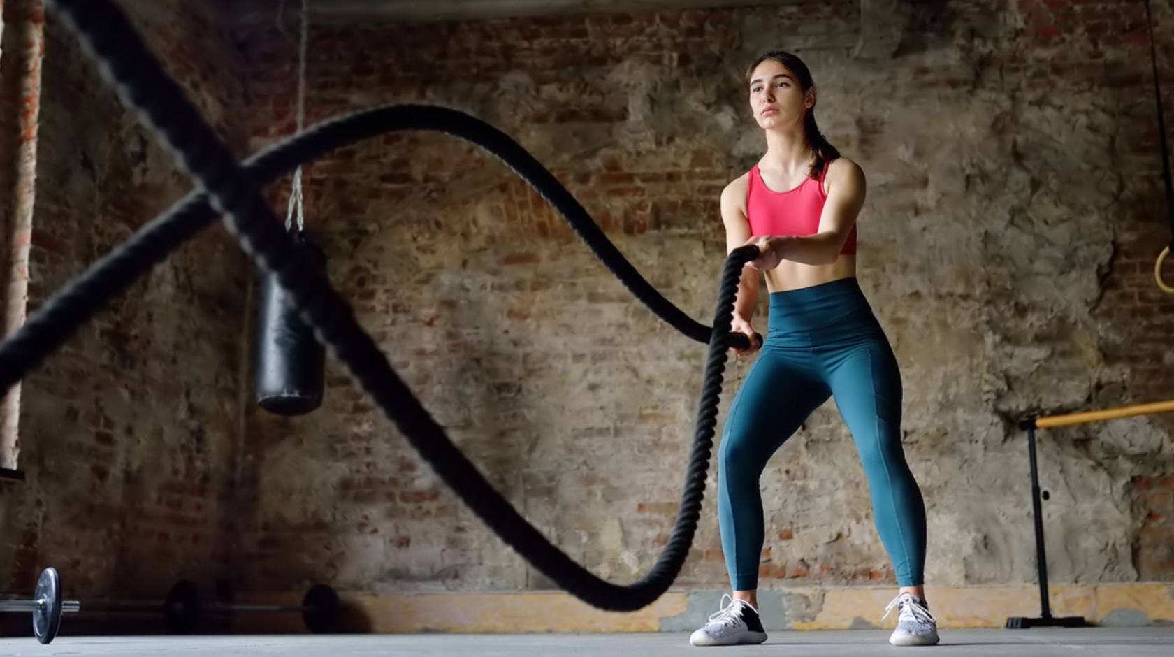 Woman in athletic attire uses battle ropes in a room with an exposed brick wall