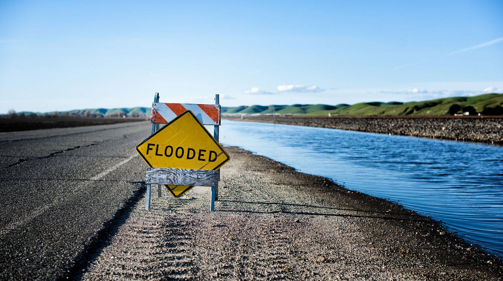 A temporary road sign reads "FLOODED", placed on a highway shoulder next to a canal, with green hills in the background