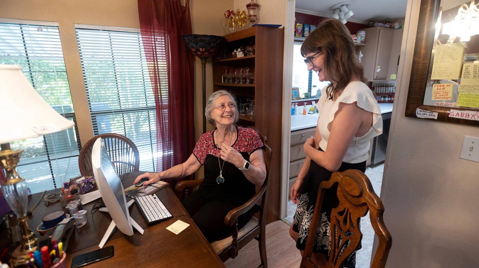 An older woman with gray hair and glasses sits at a computer at the dining room table, smiling and talking with a younger woman standing beside her