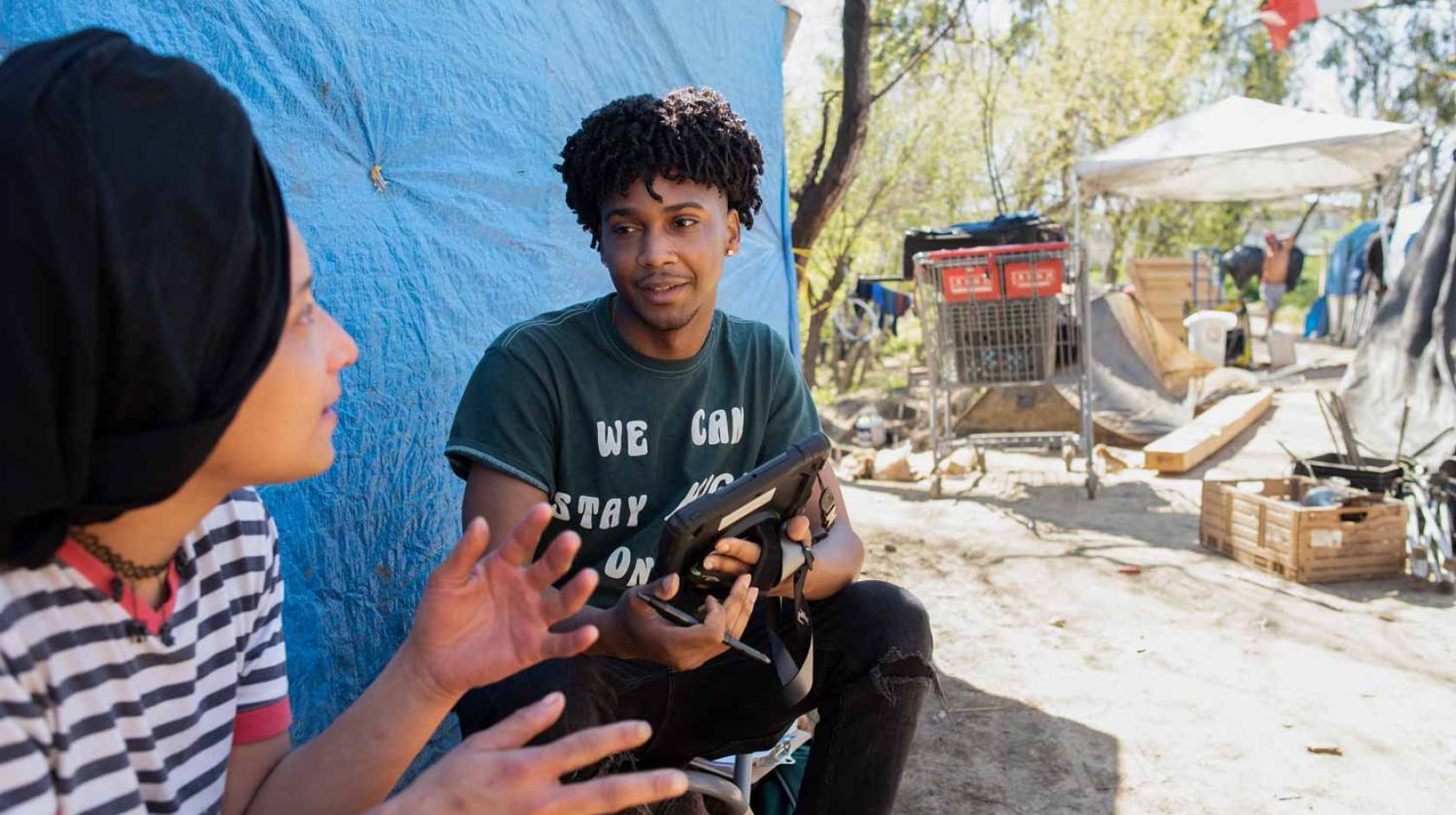 Young, attentive Black man listens to a woman in a homeless encampment and takes notes on a tablet