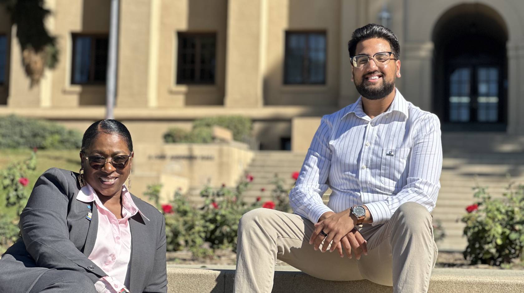 Young Black woman in a gray suit with pink blouse and sunglasses, smiling (Sonya Brooks); and young man (Josiah Beharry) with beard, glasses, khakis, dress shirt, smiling. Both seated on steps at the UC Riverside campus on a bright, sunny day.