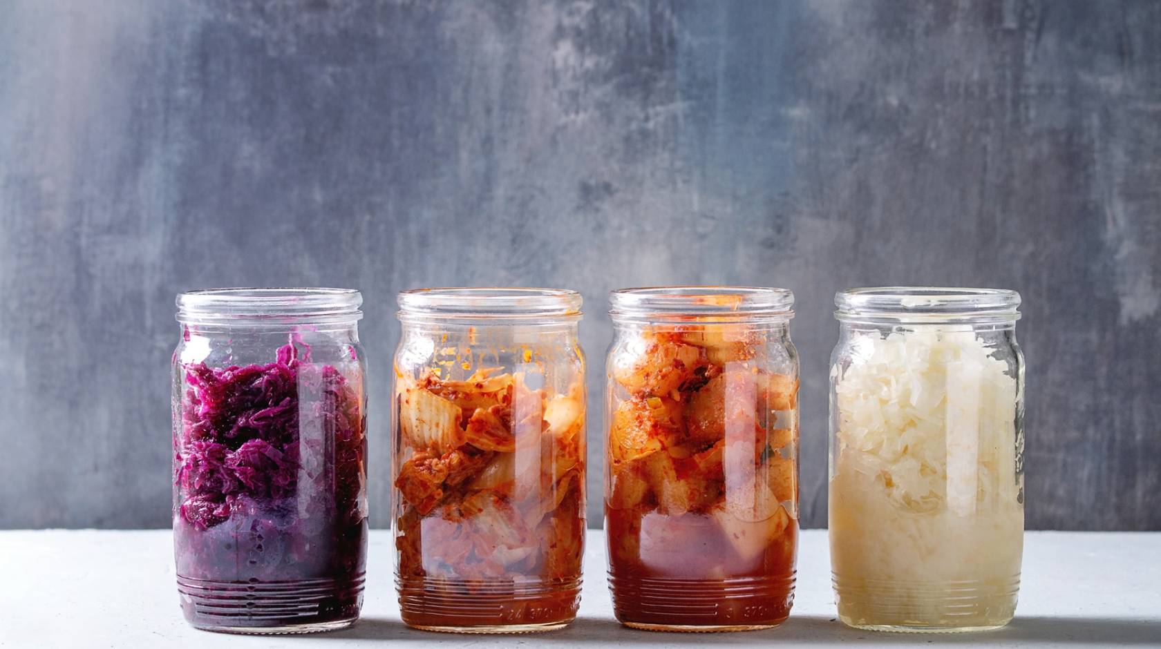 Variety of fermented food Korean traditional kimchi cabbage and radish salad, white and red sauerkraut in glass jars in row over grey blue table