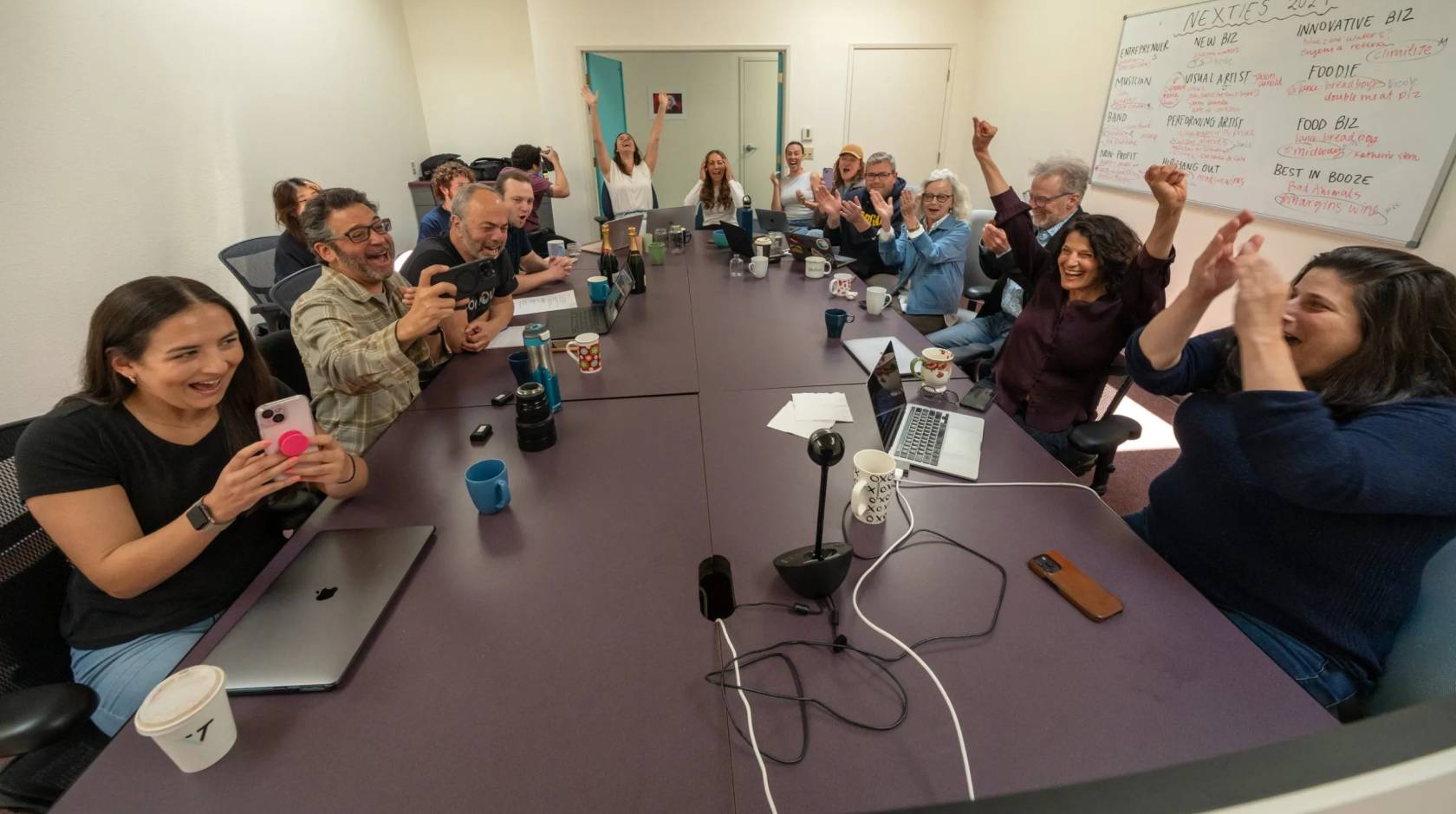 A boardroom table full of people celebrates at Pulitzer Prize win, taking selfies, raising arms, shouting, etc.