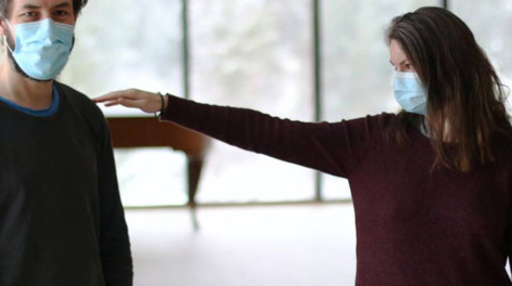 A young man and a woman wearing masks, woman reaching out to man with arm