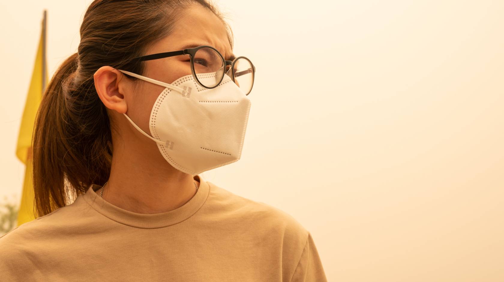 A woman with her hair pulled back wears a N95 mask, looks pensively, yellowish sky around her