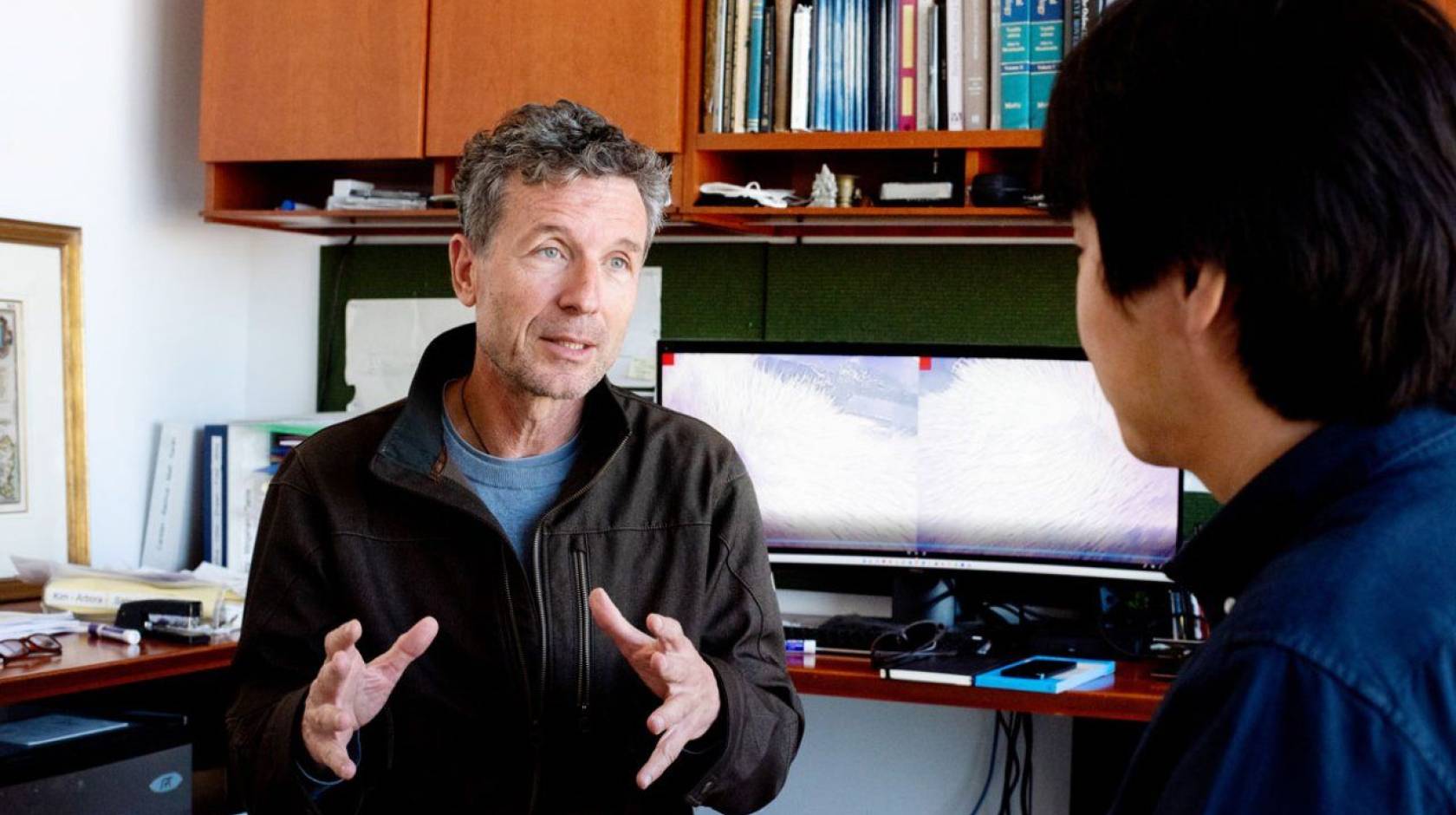 A researcher with gray hair at his desk talks to someone