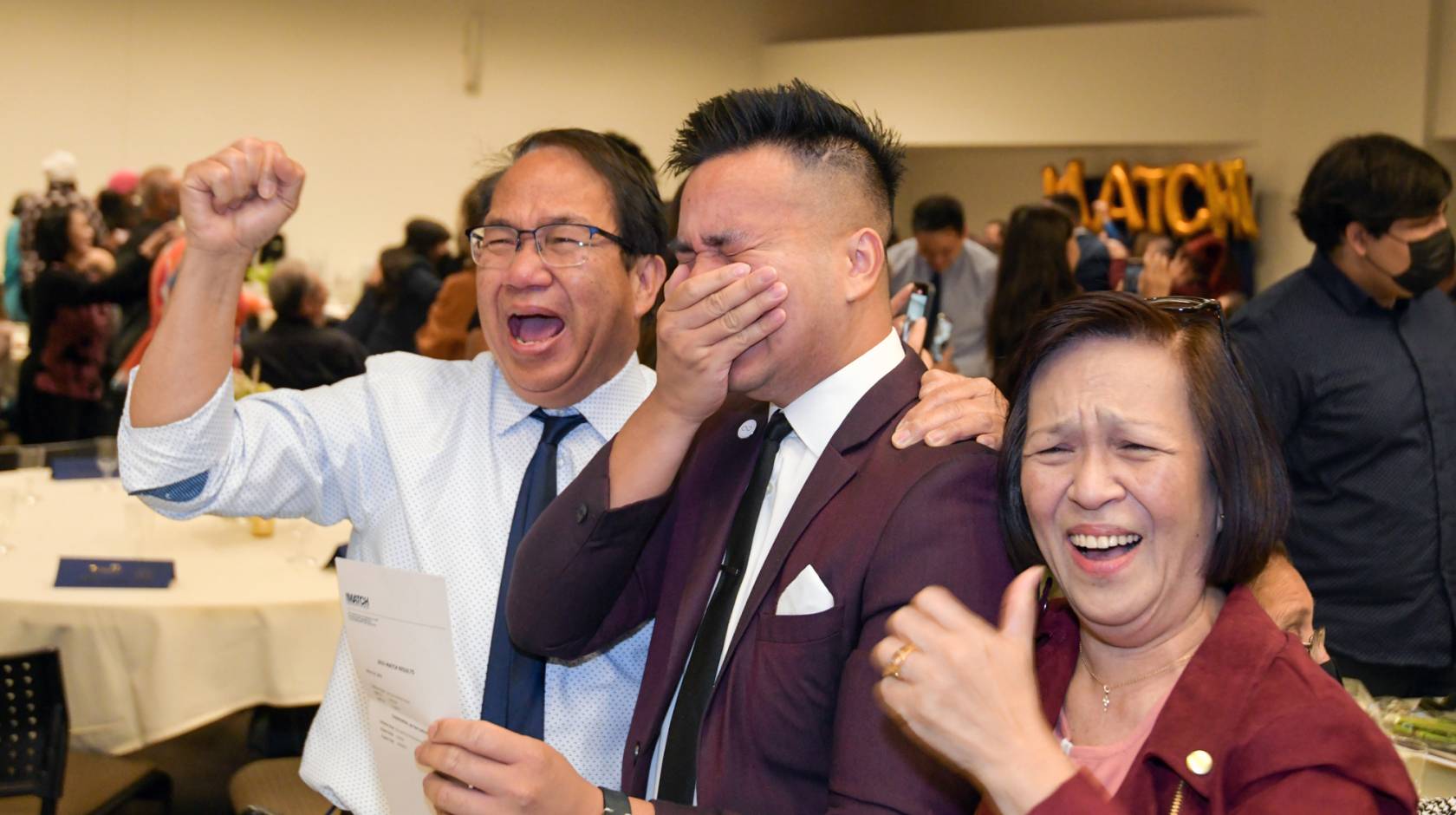A UC Riverside medical student holds his hand over his mouth in excitement, flanked by his parents, on Match Day