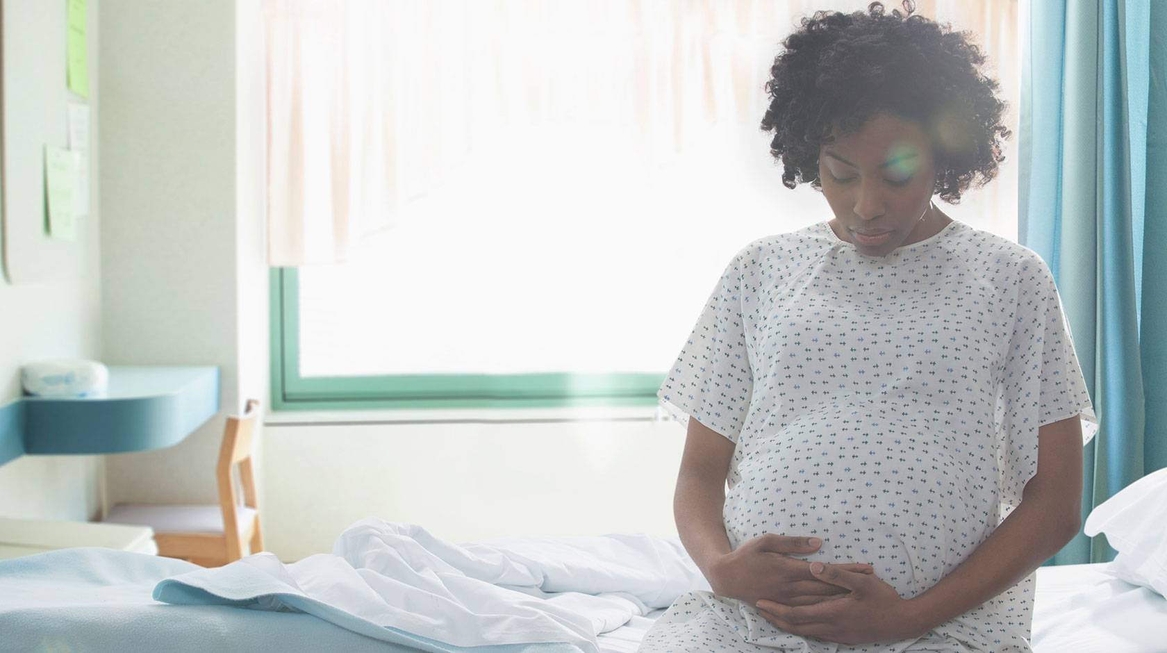 A pregnant woman sits on a hospital bed, wearing a hospital gown, holding her belly from below.