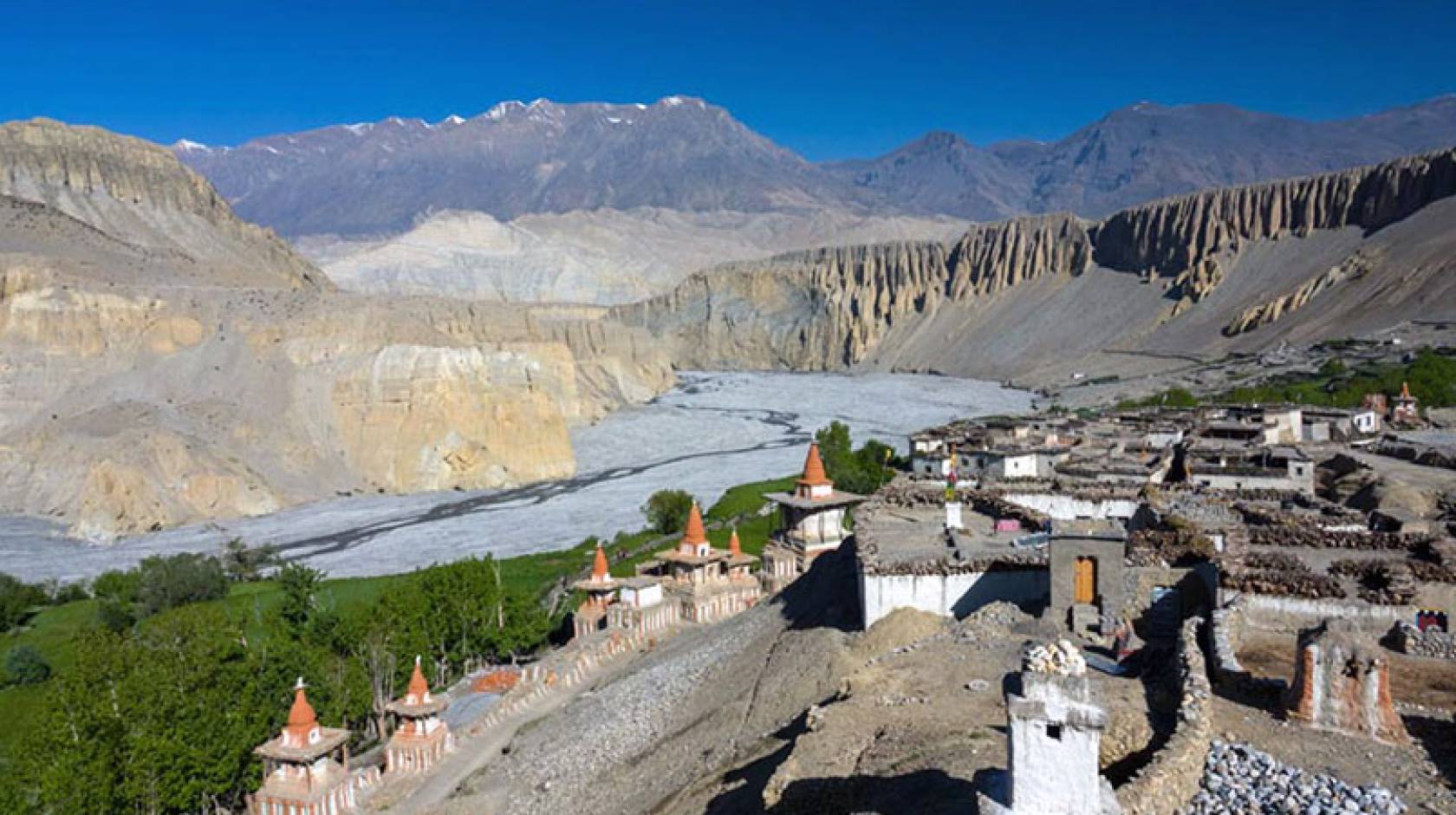 An ancient village with stone walls and buildings with pointy red roofs in a river valley with dramatic cliffs and snow-covered mountains in the background.