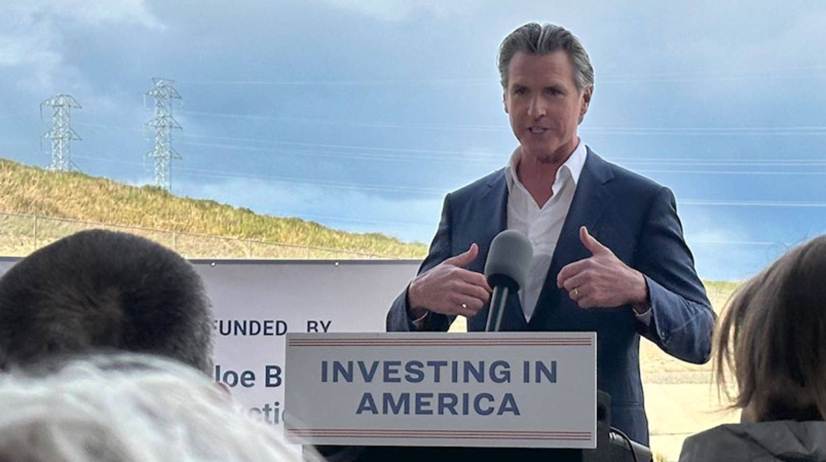 Governor Newsom speaking at a podium that says Investing in America, with countryside behind him