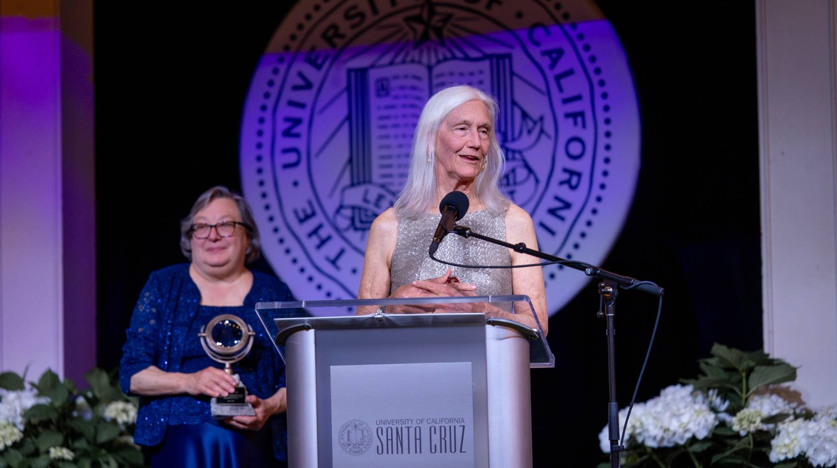 Woman with long white hair (Julie Packard) in silvery dress at a podium; woman in blue dress with short gray hair, Chancellor Larive, holding an award, stands behind