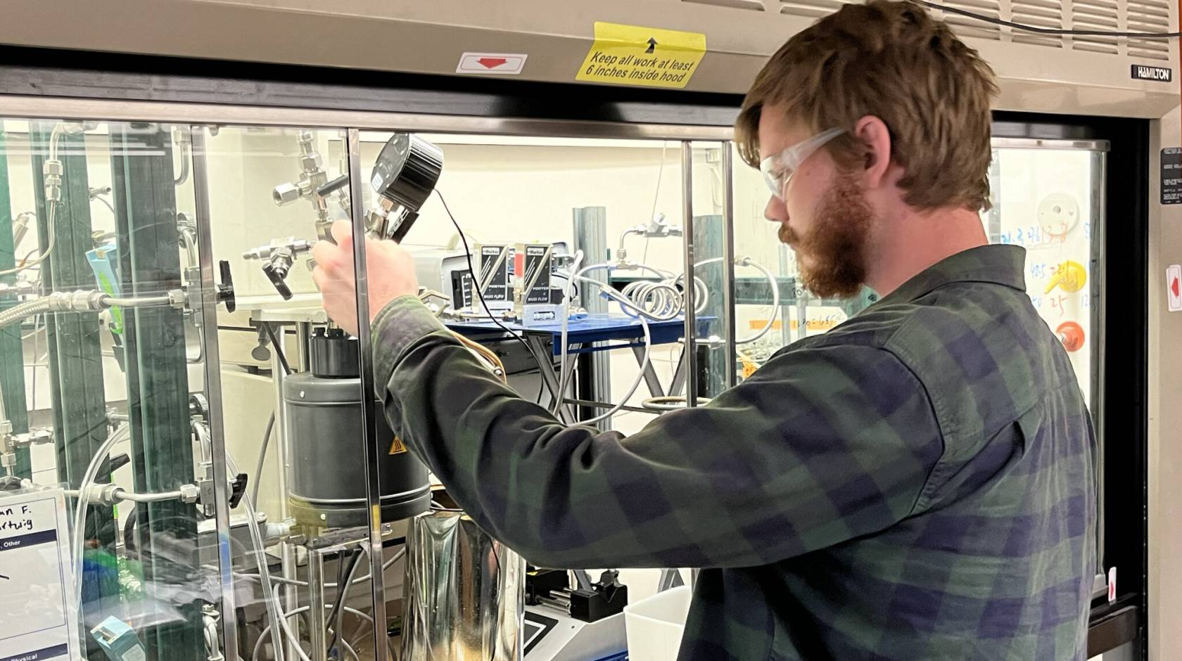 A young bearded man with red hair and protective glasses operates a small machine