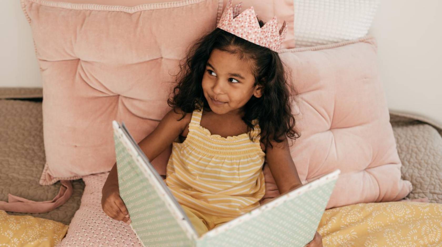 A young South Asian girl reads a book while wearing a crown and smiles