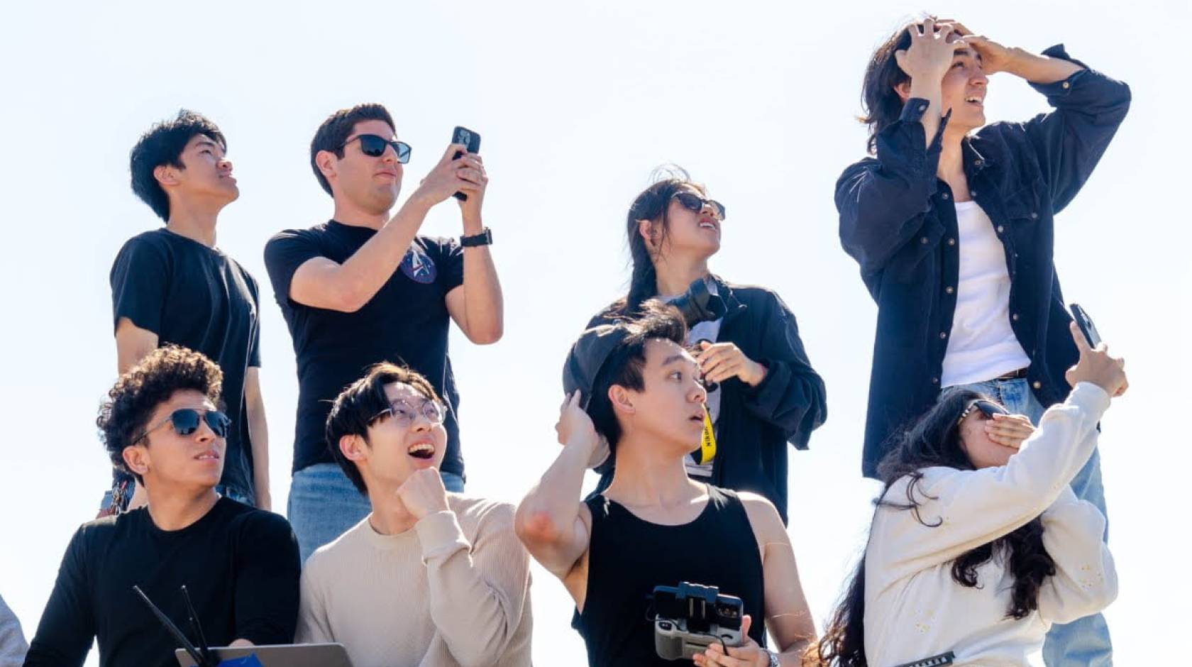 A group of eight people looking at something off camera, four standing, four seated, some gasping, a few with phones recording something, some looking shocked (presumably watching a rocket)