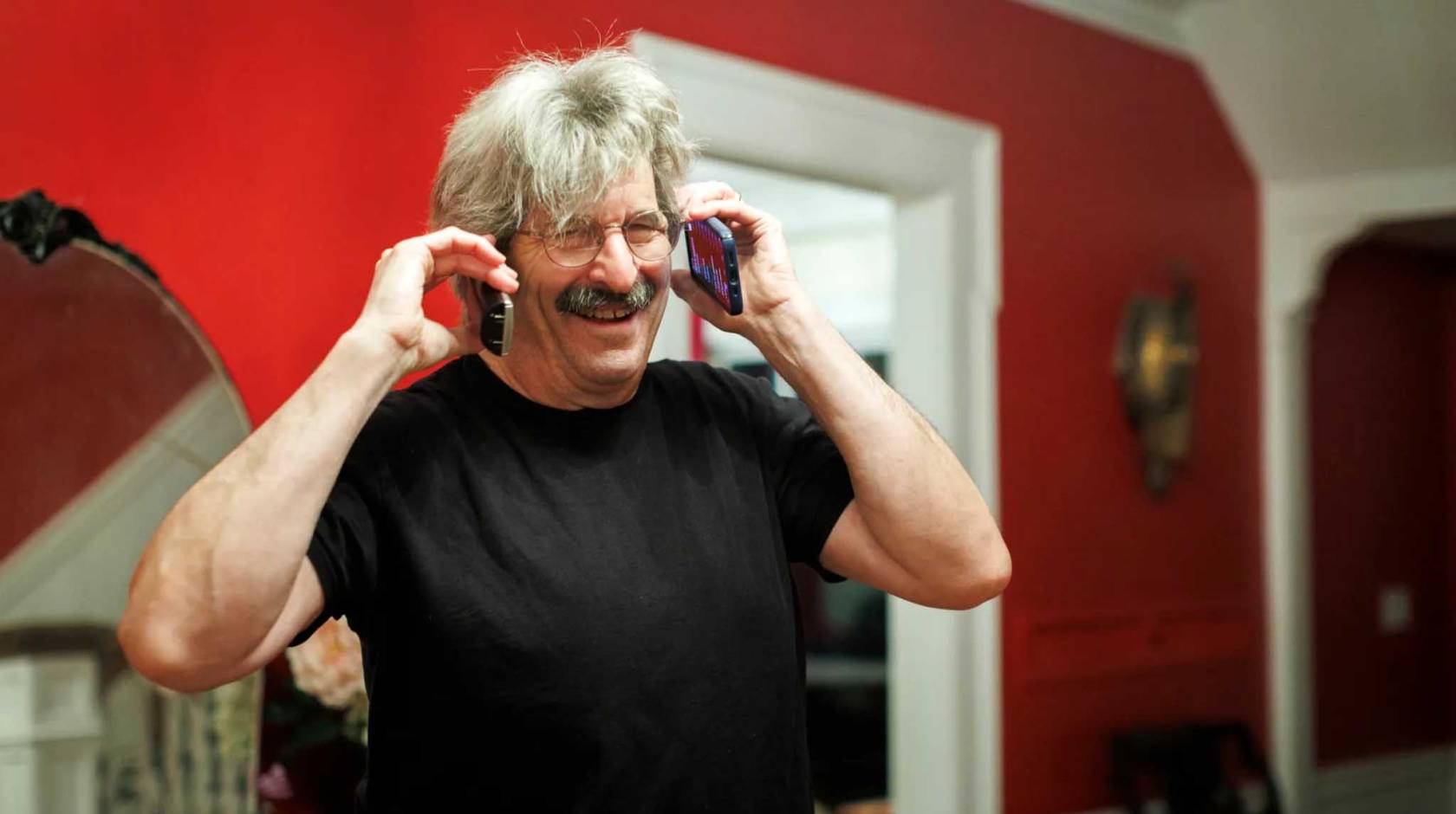 A happy man with white hair and a salt and pepper mustache answers two phones at the same time, one on each ear, laughing