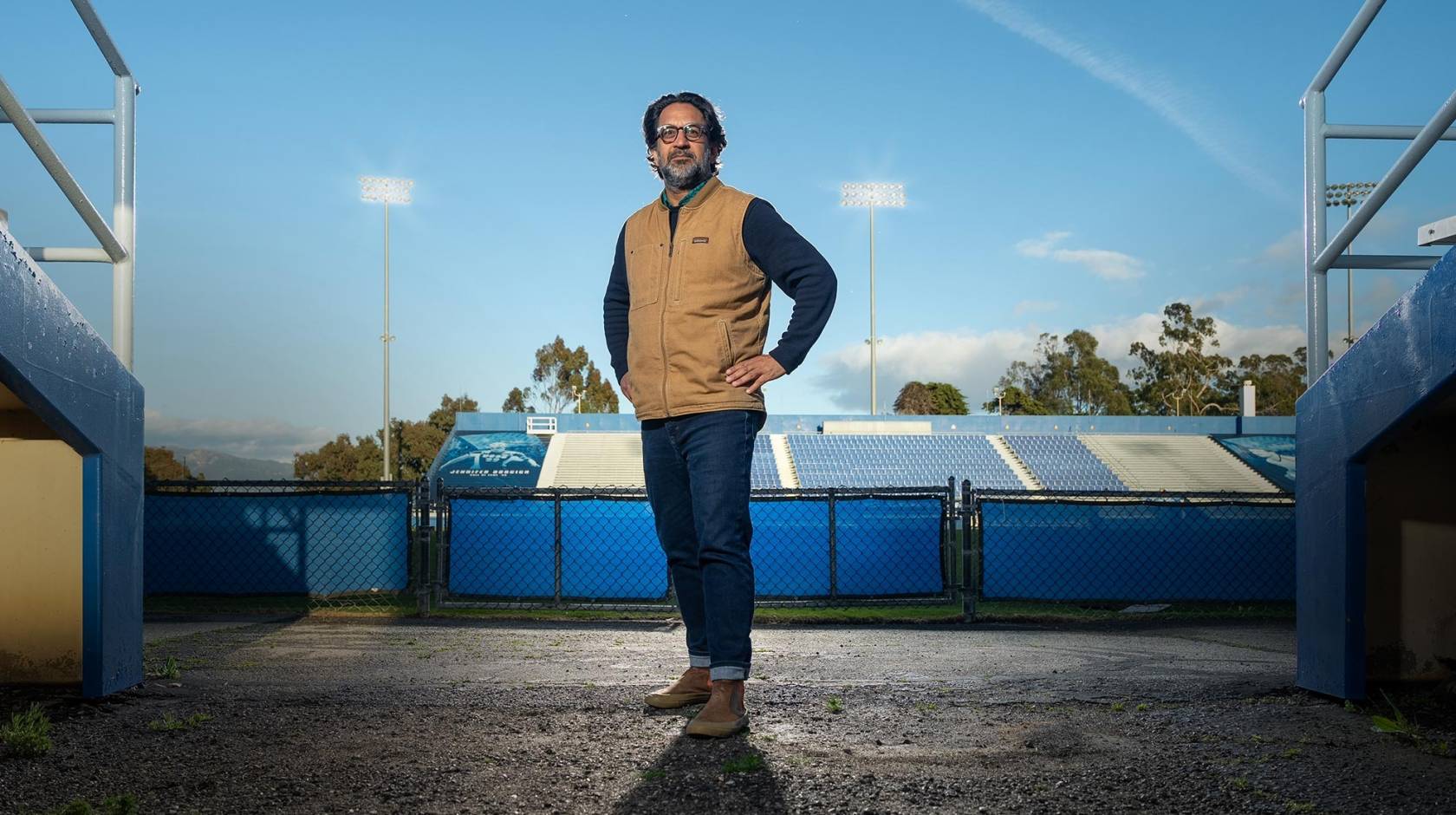 Man with salt and pepper beard stands at ground-level field entrance of a stadium, field behind him