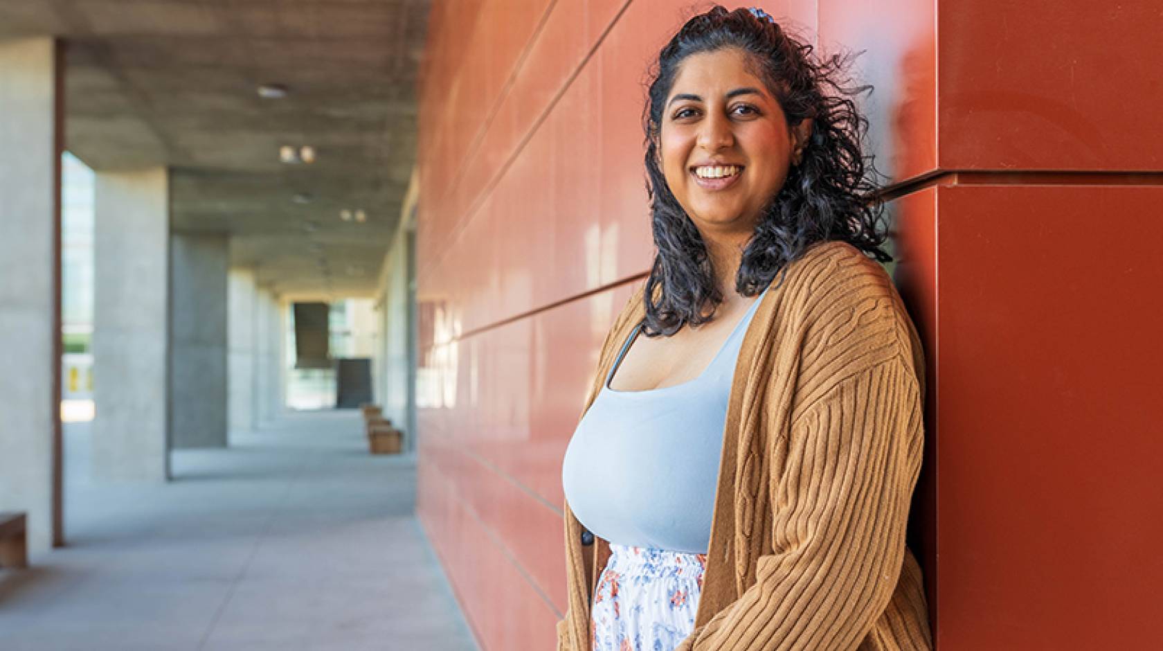 Woman with shoulder-length dark curly hair smiles while standing in an outdoor UC Merced campus corridor