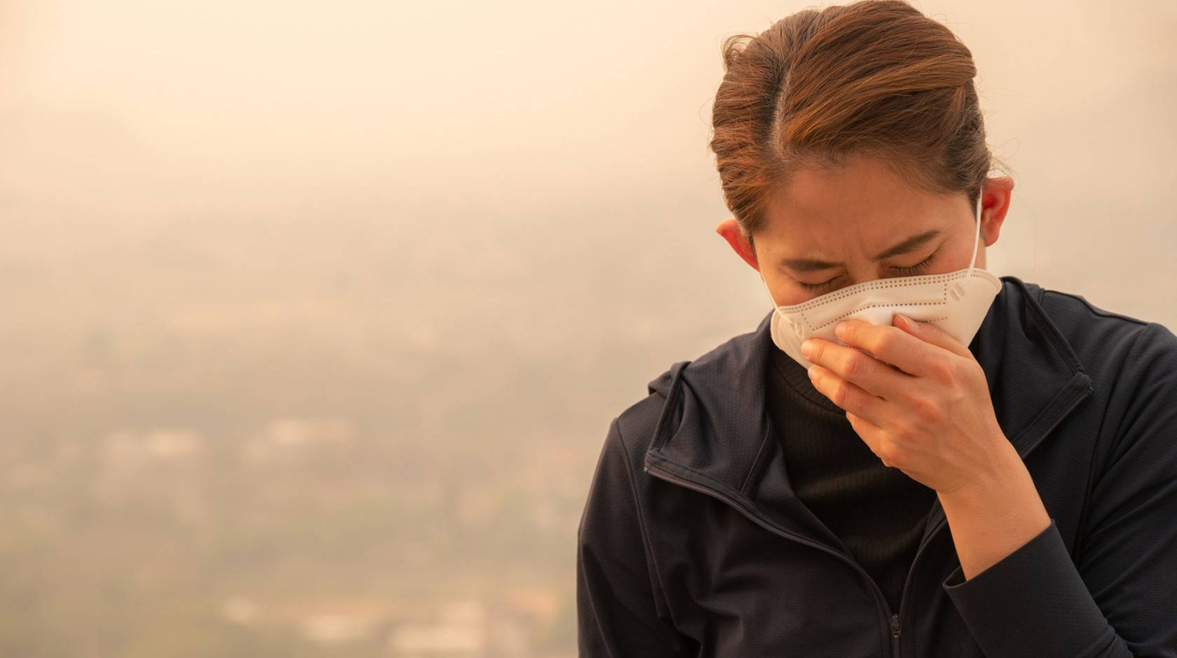 Woman wearing a face mask and coughing into her hand. They sky behind her is hazy with smoke.