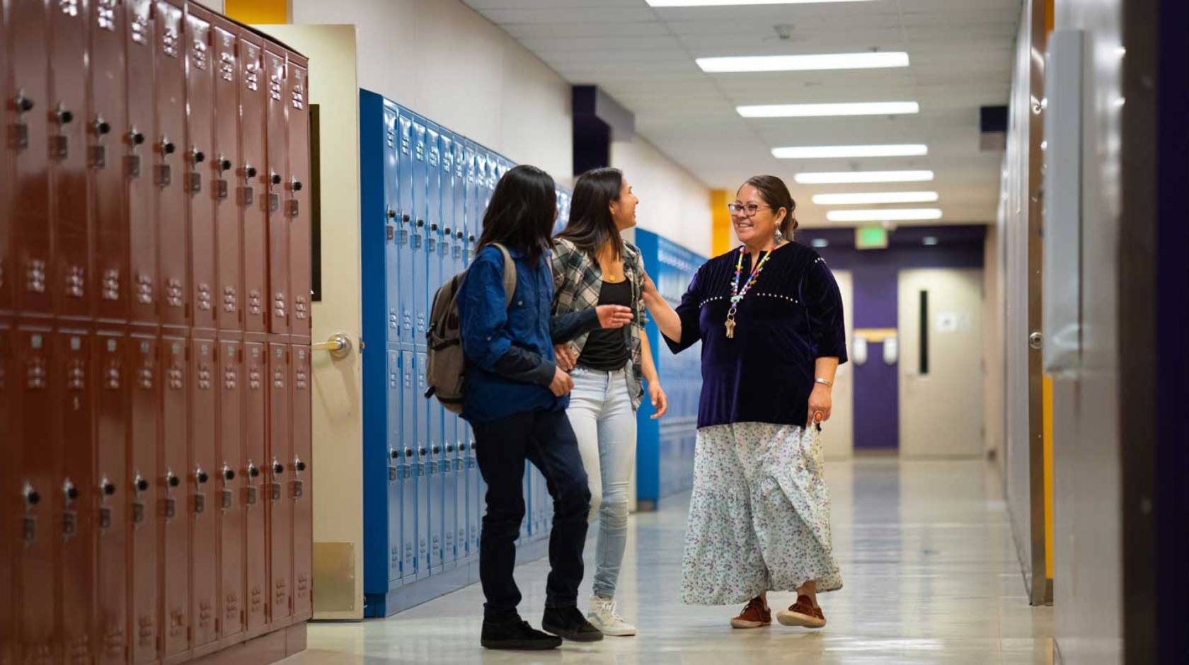 An adult talking with two high school students in a school hallway, with rows of lockers in the background