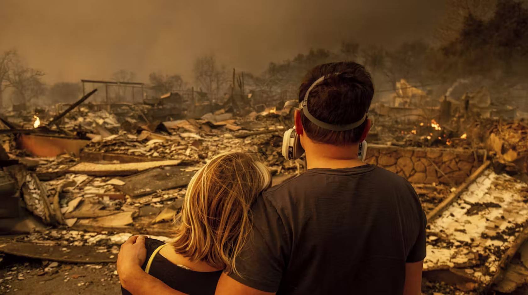 A young man in a mask and woman, photographed from behind, look on at an obliterated house with smoke all around