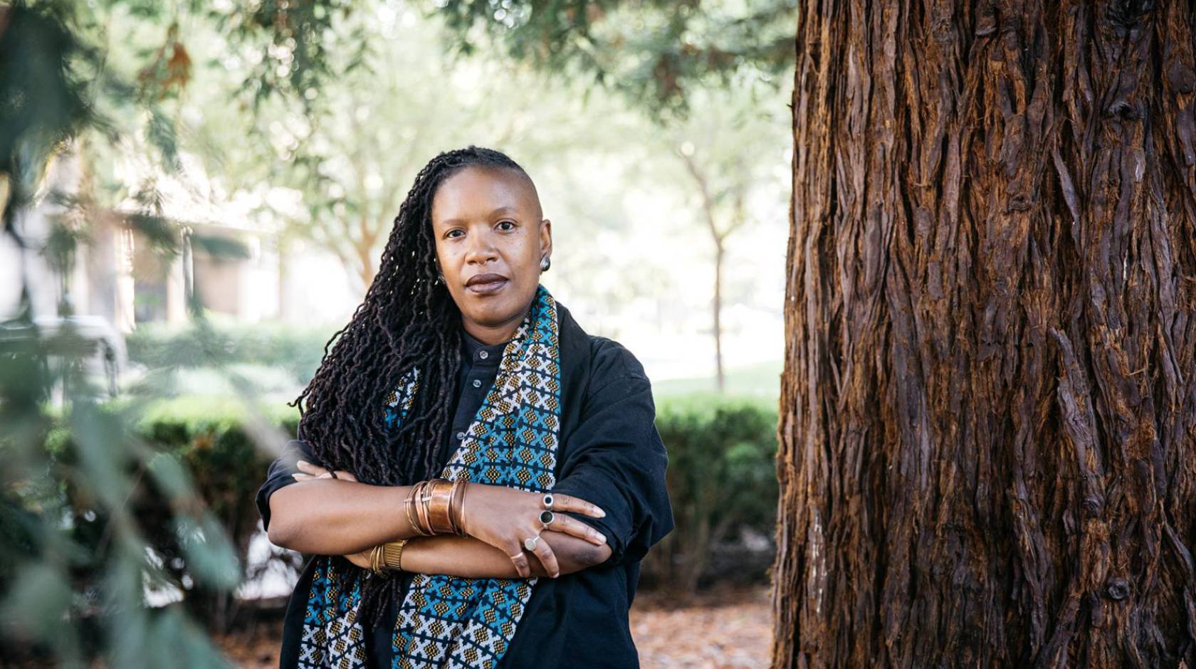 E. Tendayi Achiume, African American woman, poses outside, arms crossed, next to a redwood