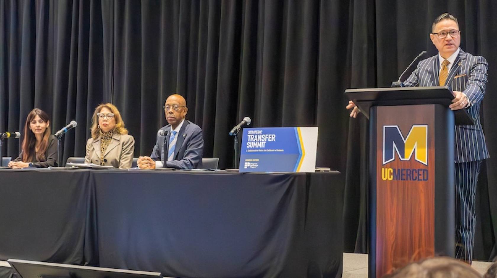 At a table, three people sit in front of microphones, from left: California Community Colleges Chancellor Sonya Christian, California State University Chancellor Mildred Garcia, and UC President Michael Drake; at a lectern, UC Merced Chancellor Juan Sánchez Muñoz stands. Audience is visible a bit in the front of the photo 