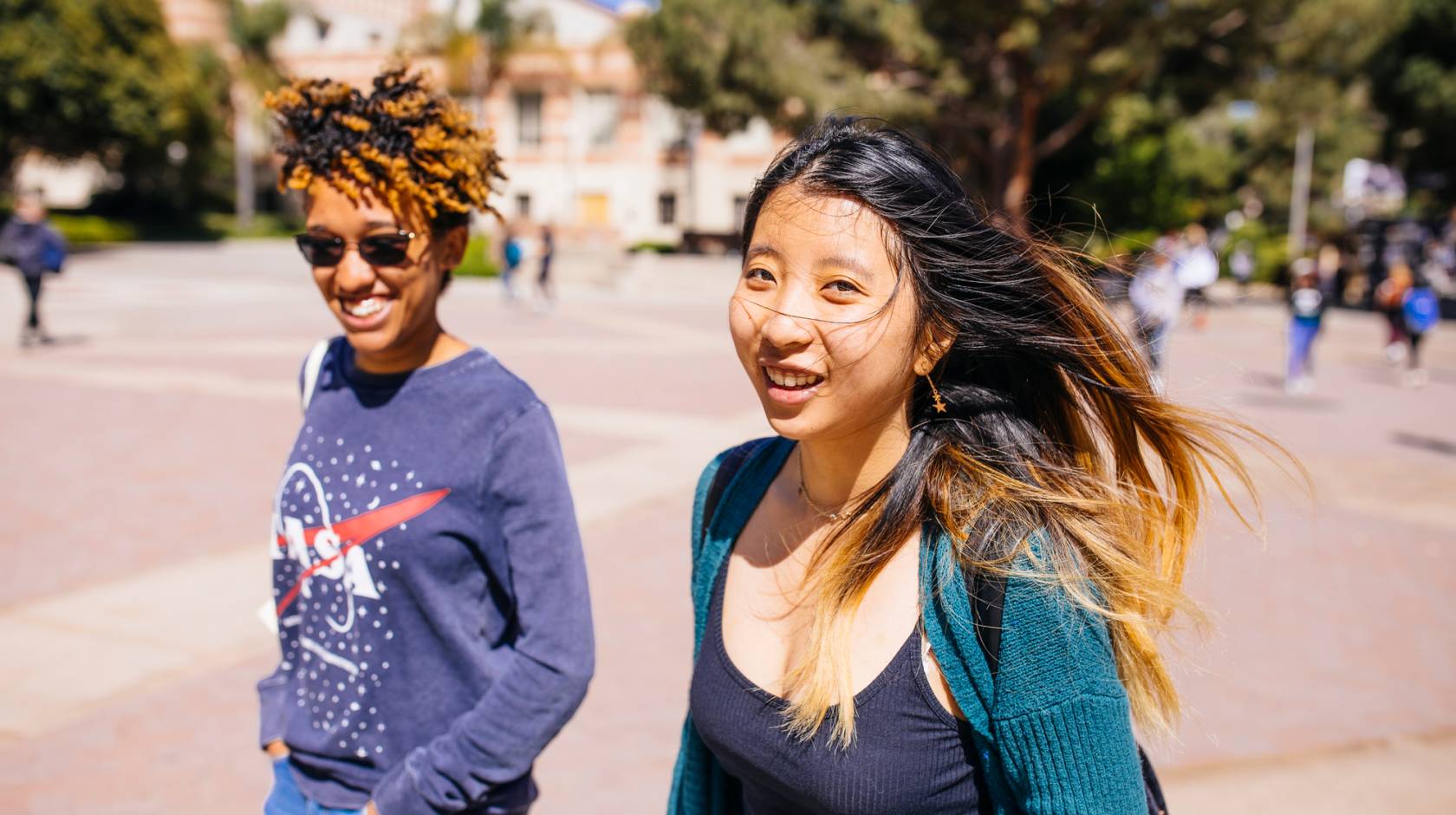 A young Black woman with a NASA sweatshirt and a young Asian woman with a green cardigan walk along a campus smiling