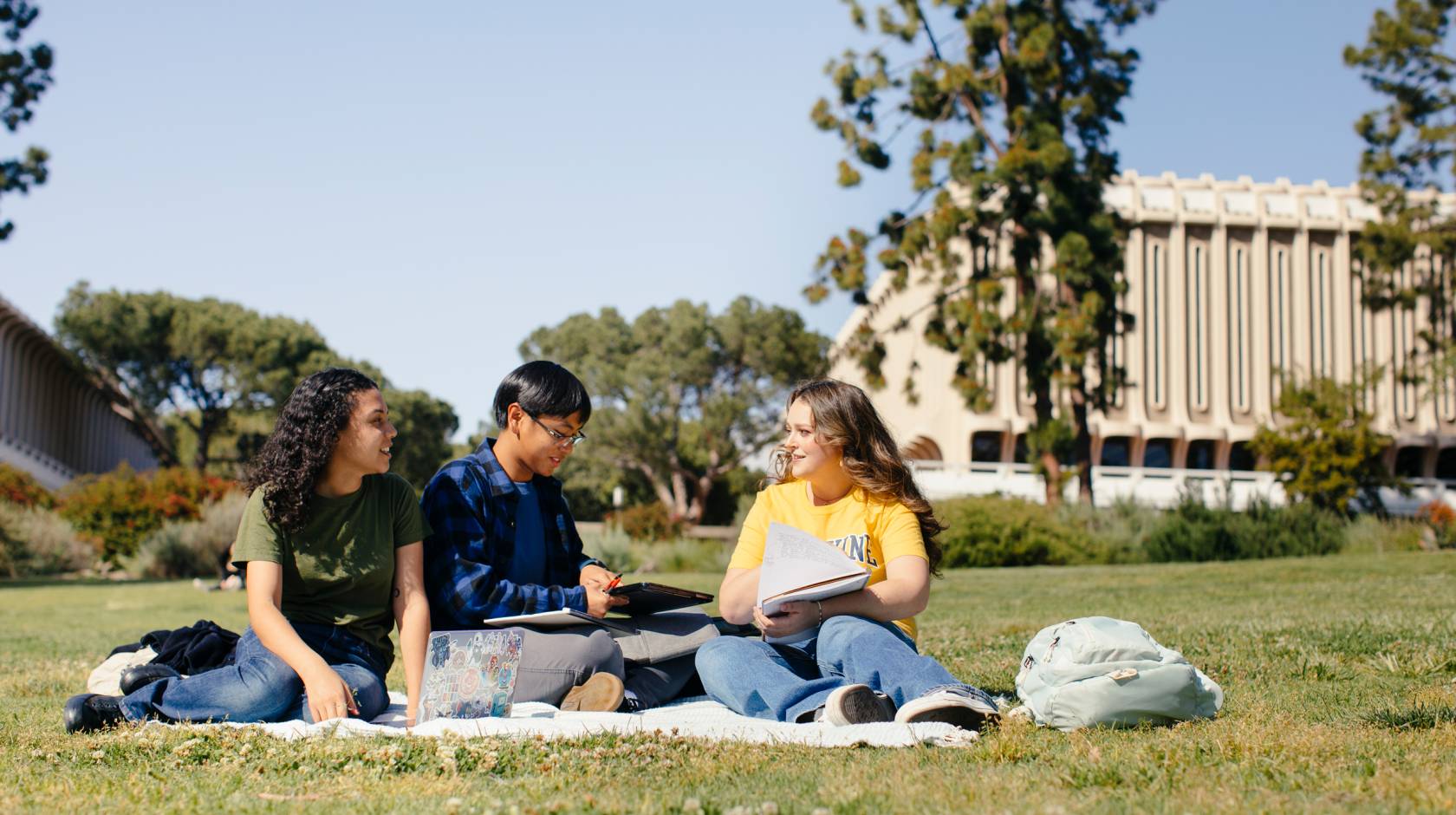 Three students study together on a blanket on the lawn of the UC Irvine campus, tall trees and buildings visible in the background