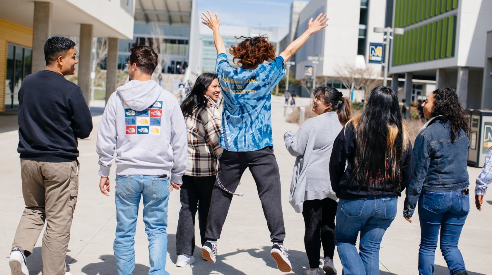 Viewed from behind, a diverse group of students walks along the UC Merced campus, the center student jumping exuberantly