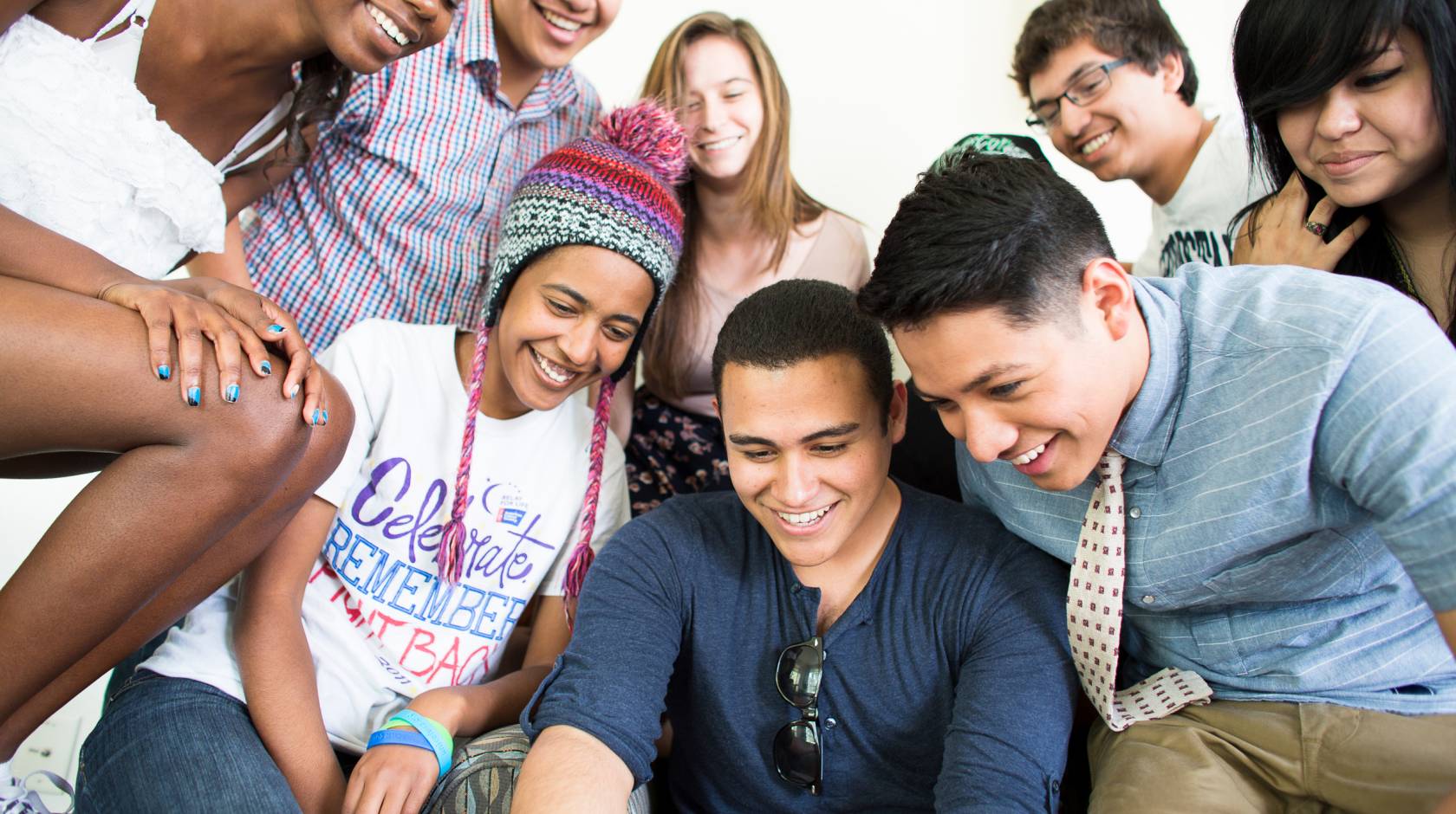 A diverse group of students gather together to look down at something out of frame, like a computer