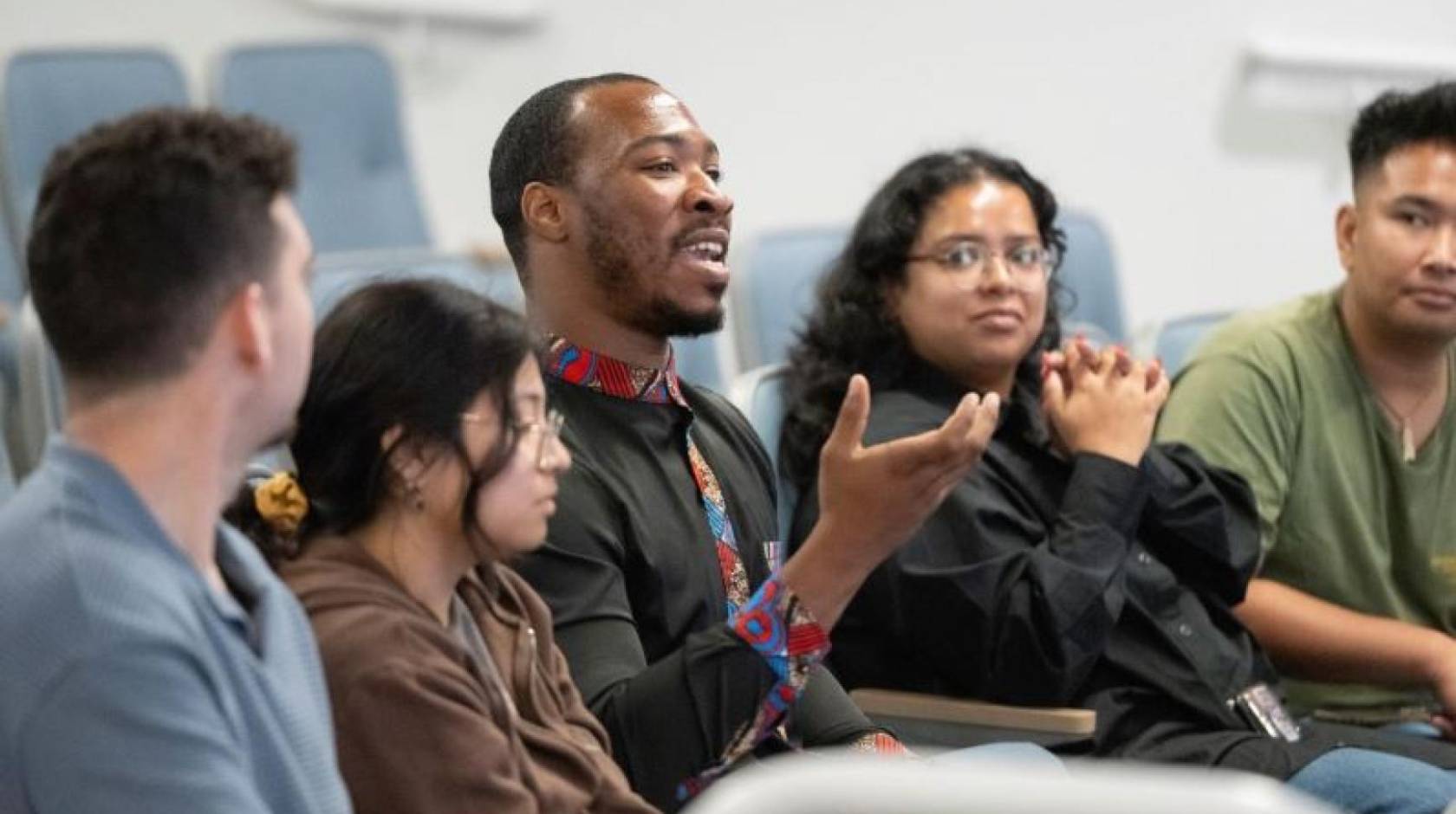 Five students in a row at a lecture hall. The male student in the middle speaks and gesticulates. 