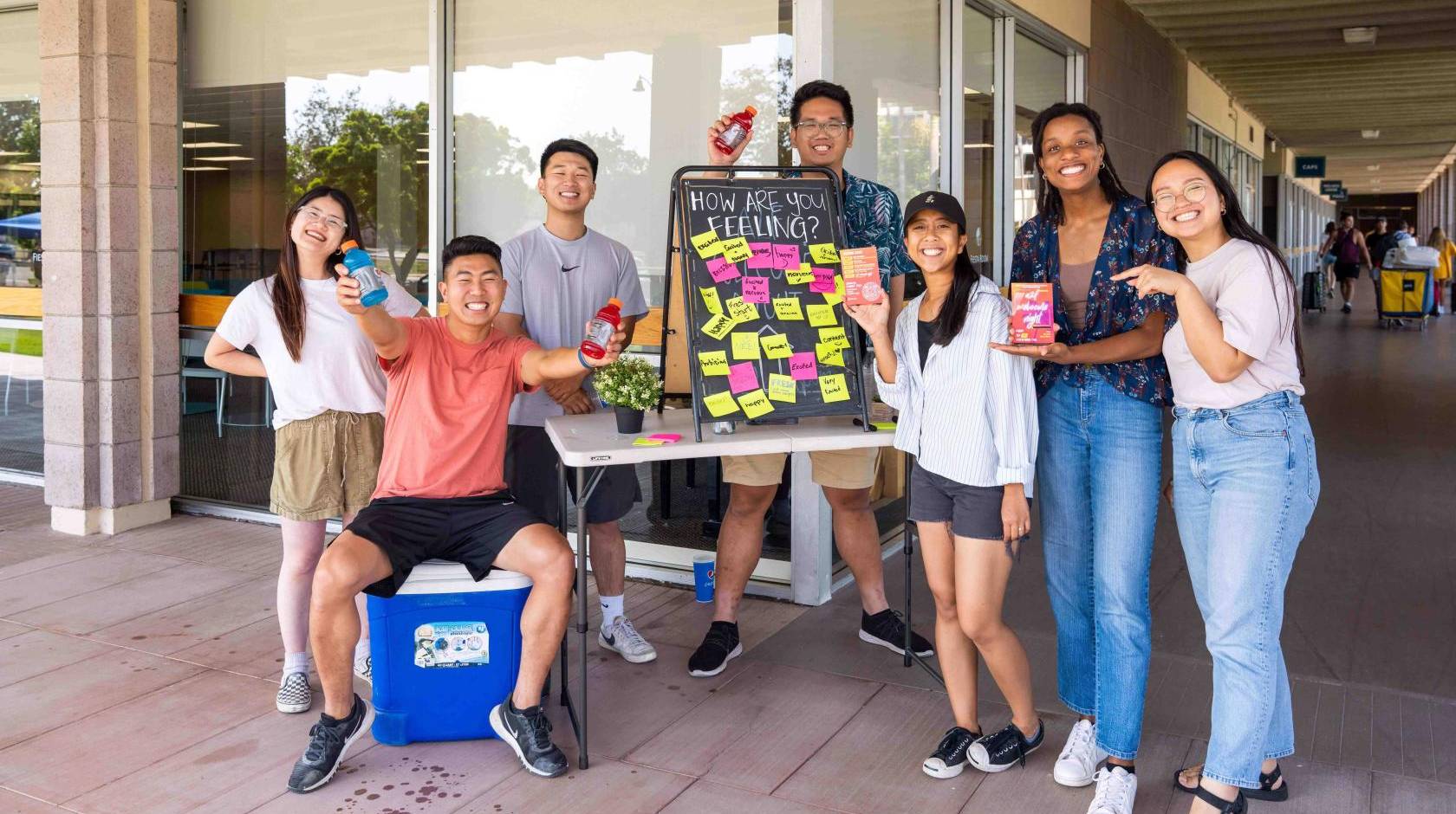 A group of smiling students in front of a dorm with bottles of water and a board that says How Are You Feeling and a number of responses