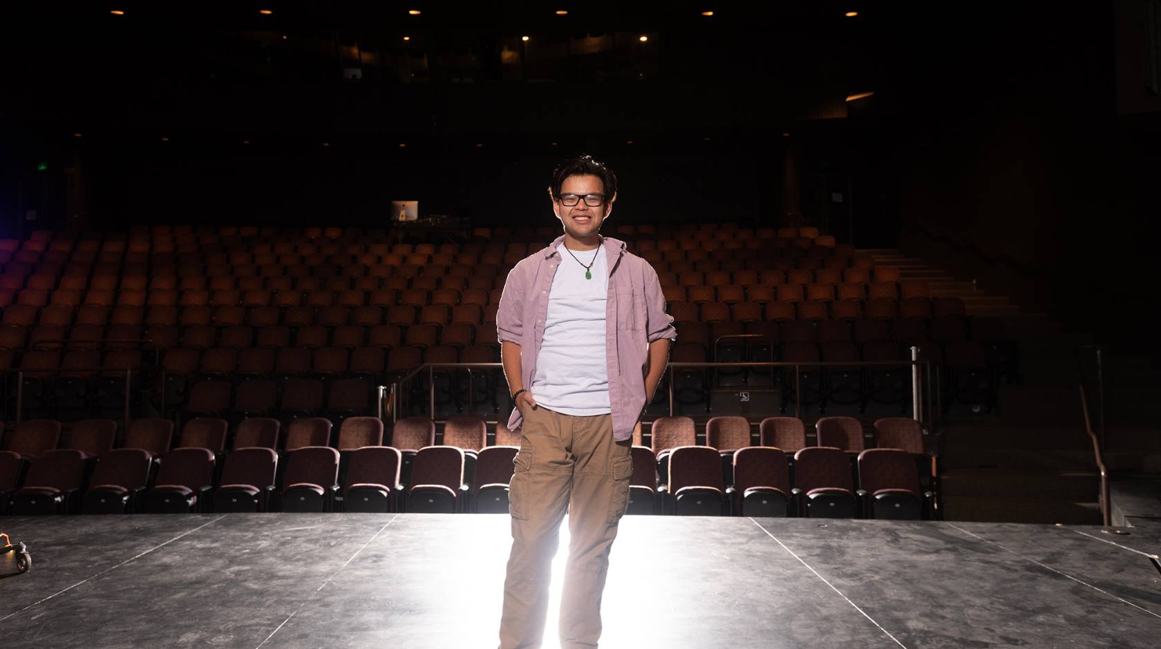 A smiling man standing on an empty theater stage under dramatic lighting