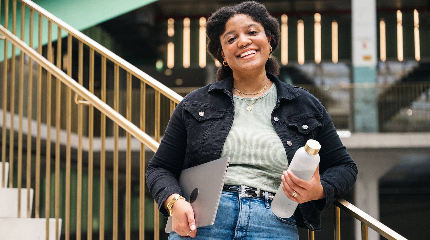 A college student smiles for the camera on a staircase in a university building, holding a reusable water bottle and a laptop