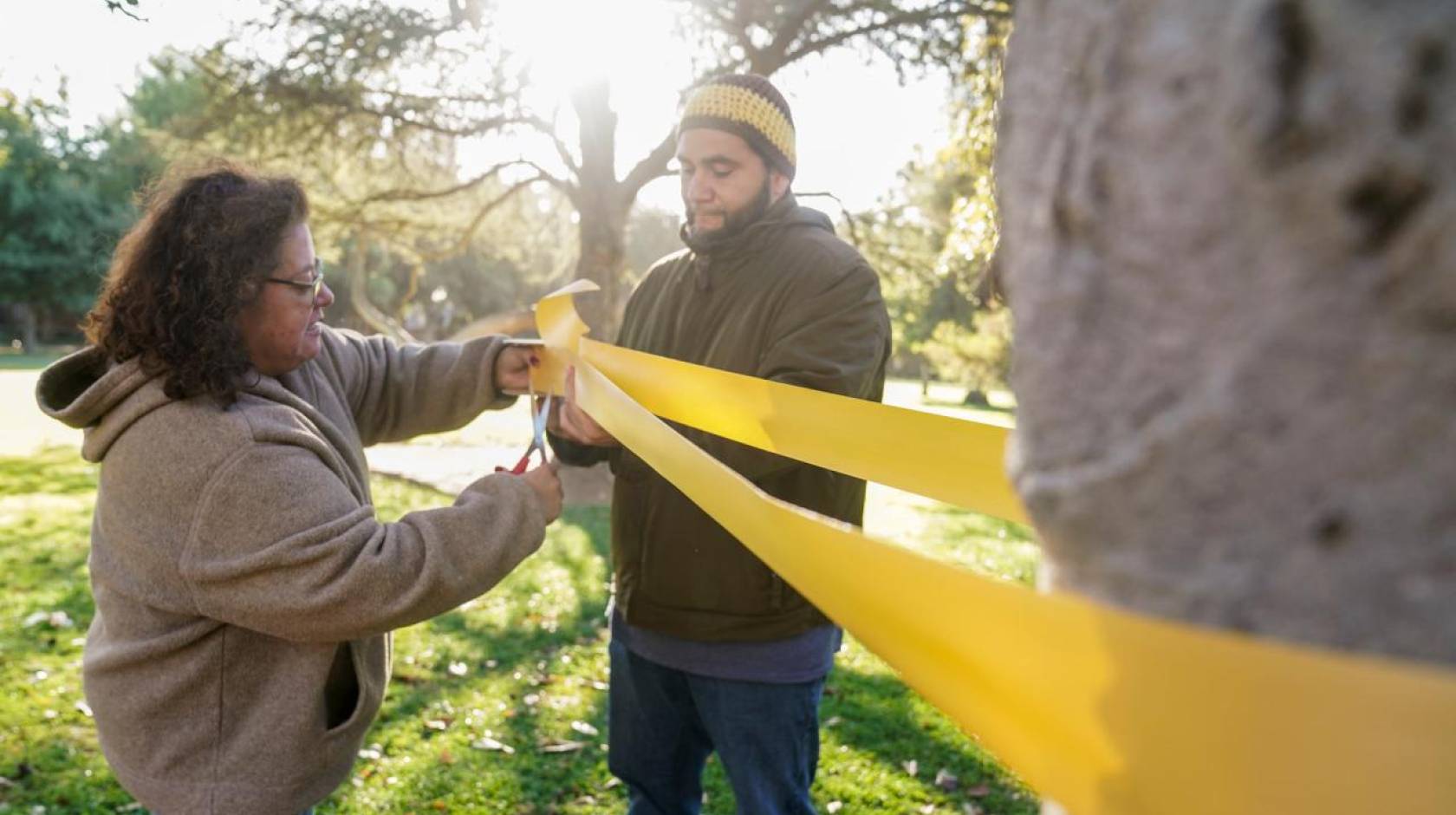 A woman cuts a long yellow ribbon tied to a tree a man is holding