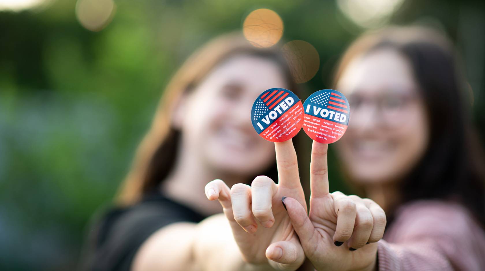 Two young women, mostly out of focus, hold up I Voted stickers to the camera, smilling